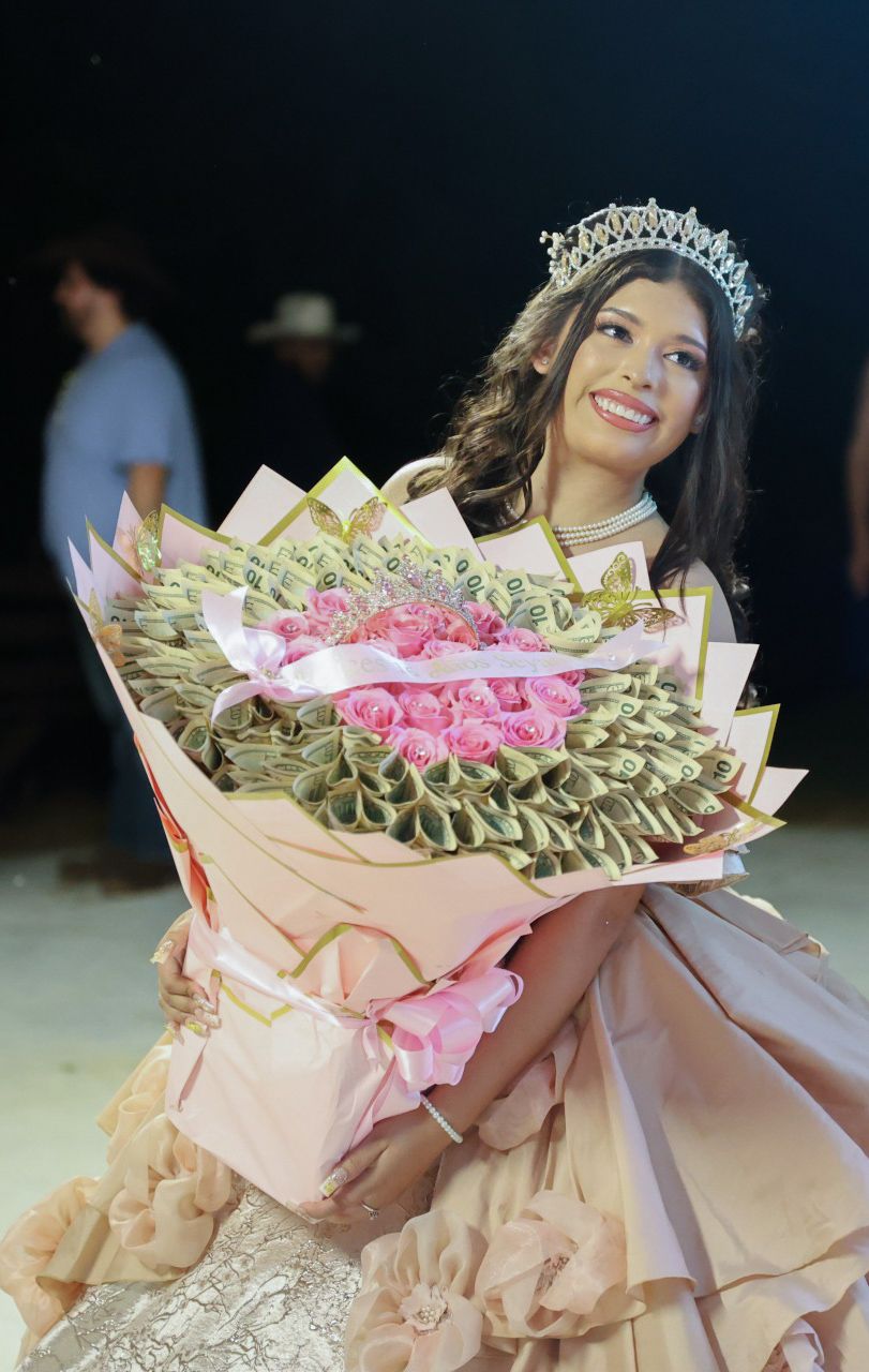 Smiling quinceañera girl wearing a tiara and pink gown holding a large bouquet of roses and money flowers.