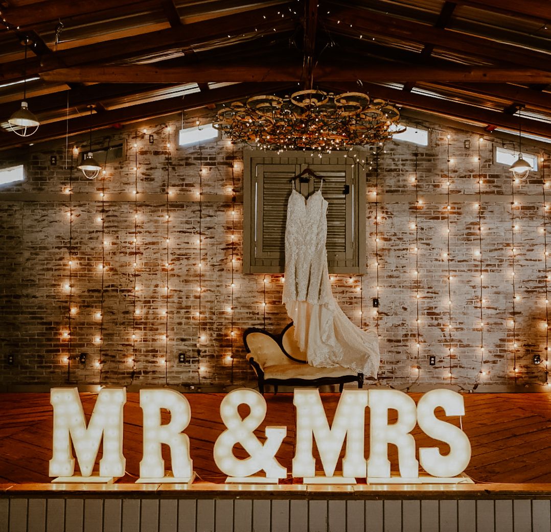 Large illuminated MR & MRS marquee letters at a rustic wedding venue with fairy lights and a bridal gown displayed on the wall.