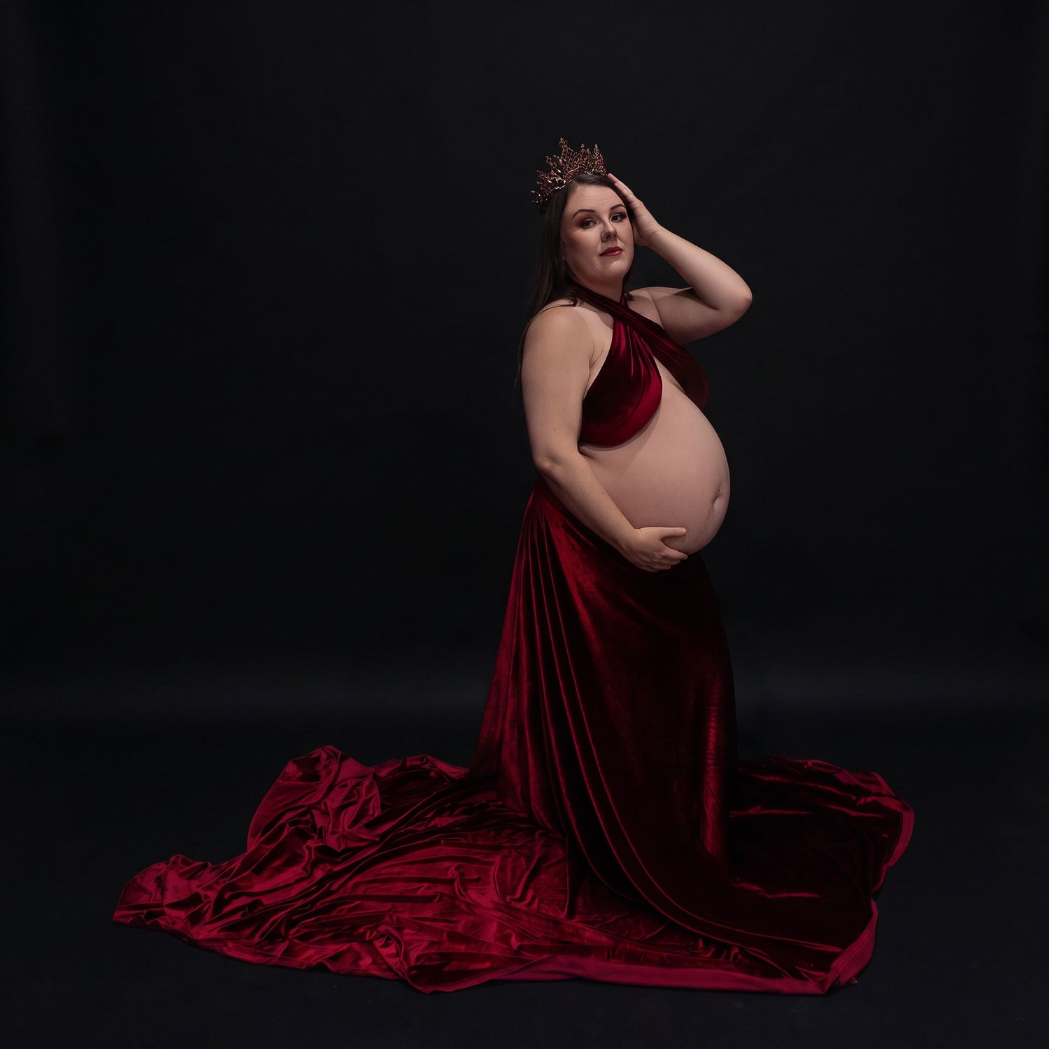 Pregnant woman in red velvet gown and crown poses elegantly against dark background in maternity photo shoot.