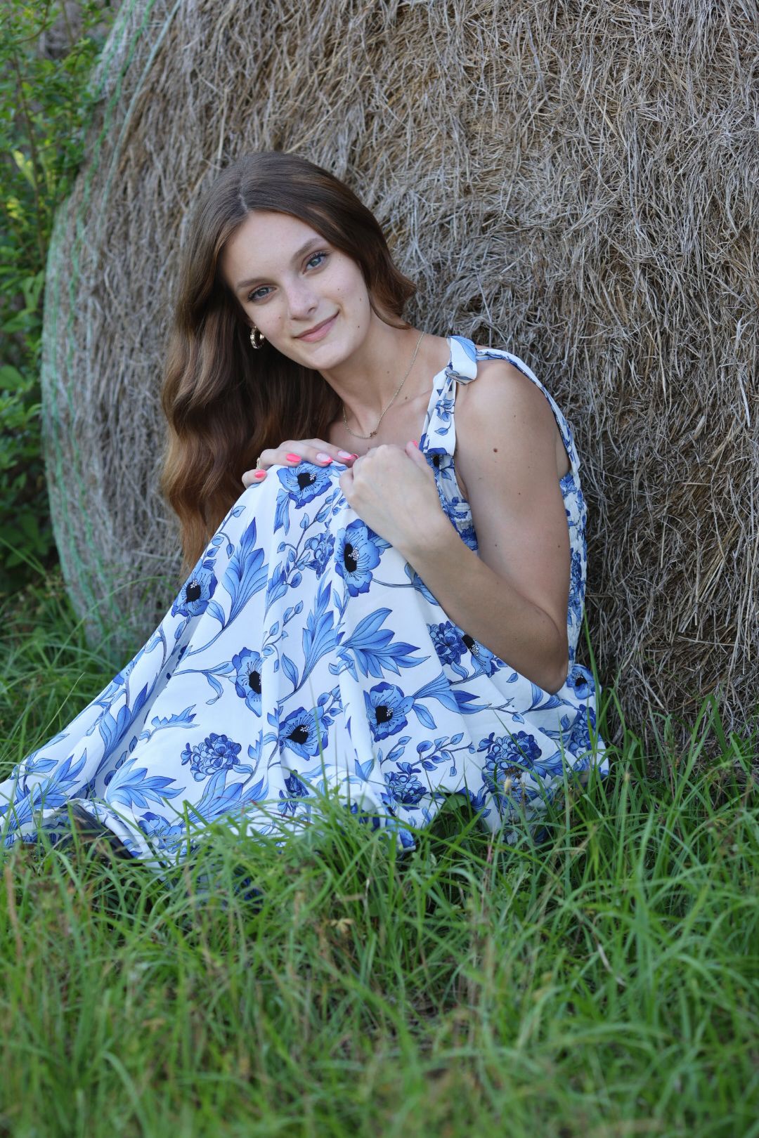 Young woman in blue floral dress sitting against a hay bale in a green field, smiling warmly.