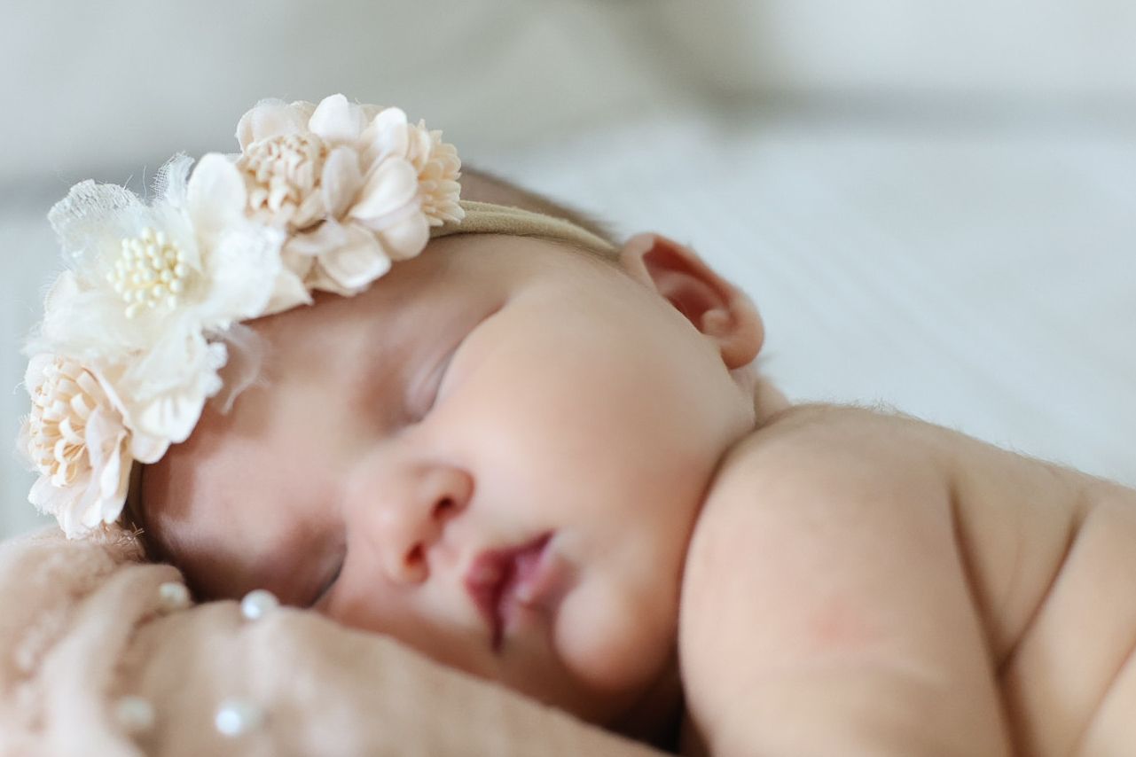 A sleeping baby wearing a delicate floral headband made of cream-colored flowers rests peacefully on soft bedding.