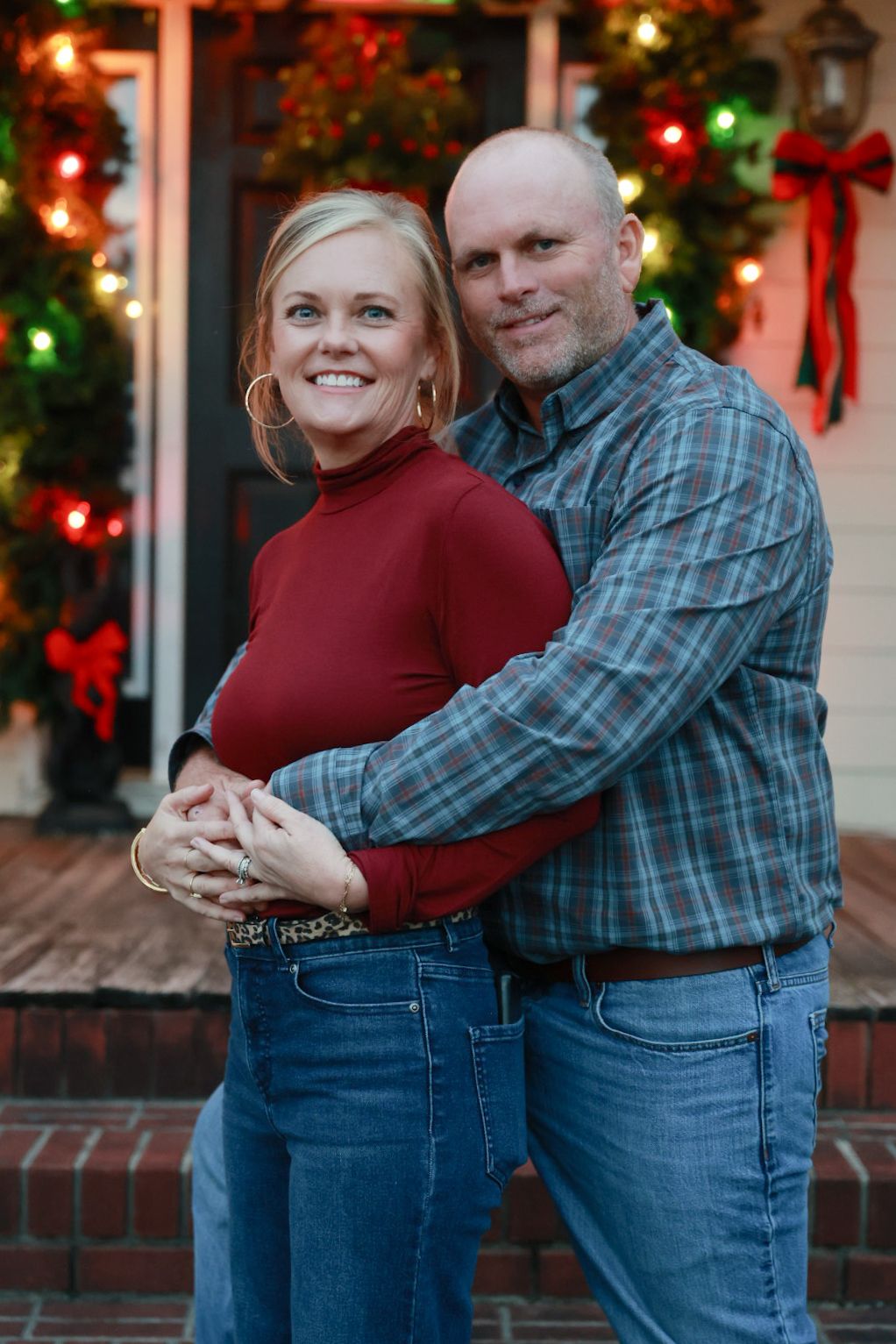 A couple embraces in front of a festive holiday display with red and green Christmas lights and decorative bows.