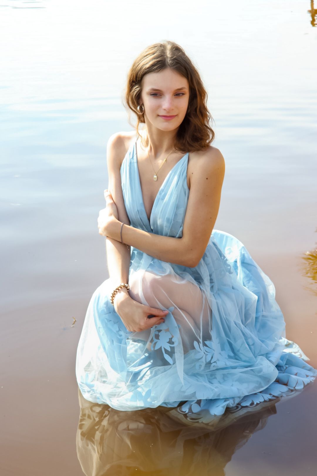 Young woman in a light blue floral gown sitting gracefully in shallow water, smiling softly.