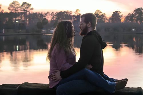 Couple sitting close together on a dock at sunset, sharing a romantic moment by a tranquil lake.