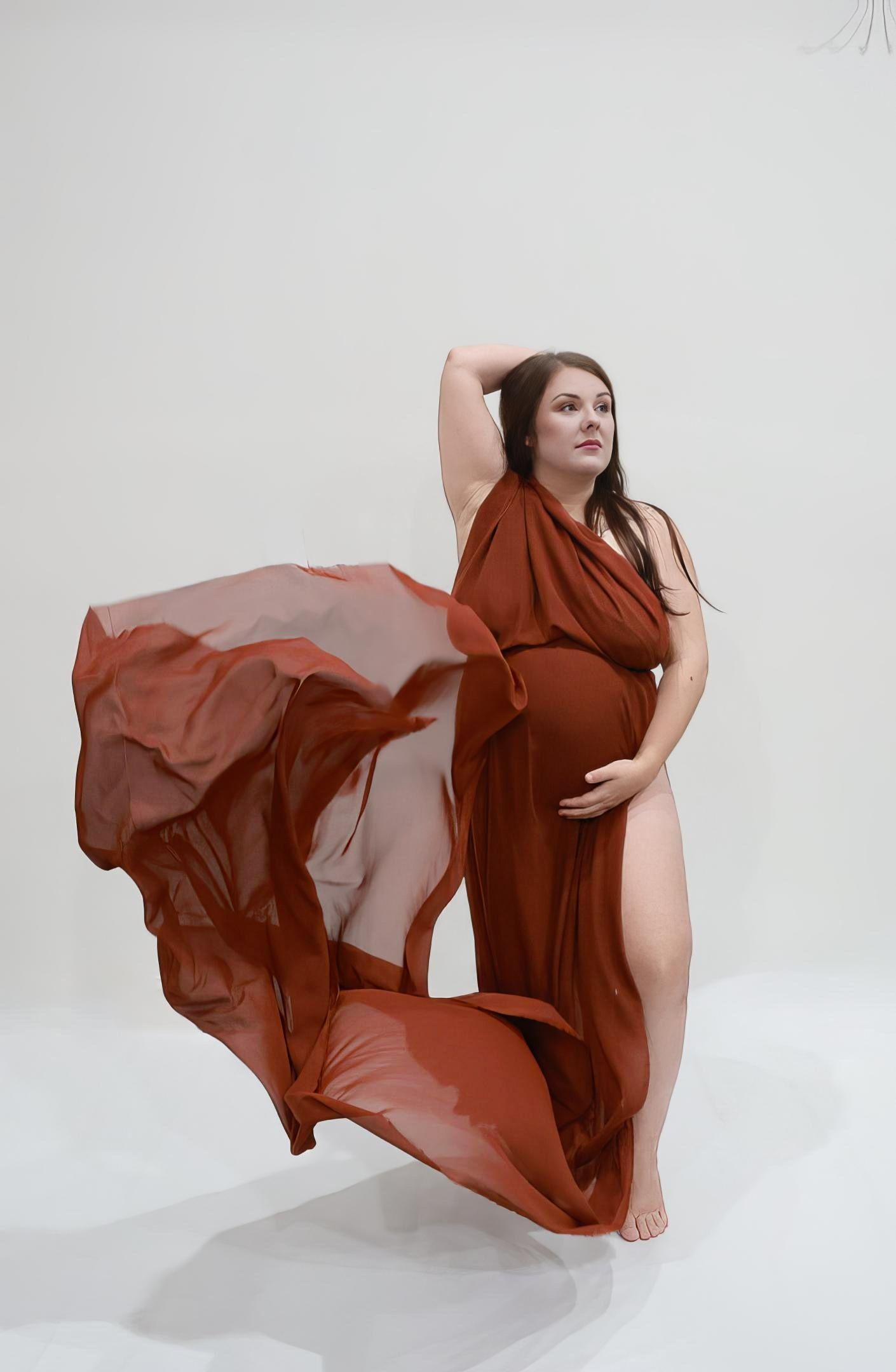 Pregnant woman draped in flowing rust-colored fabric poses gracefully in a bright white studio for a maternity photo shoot.