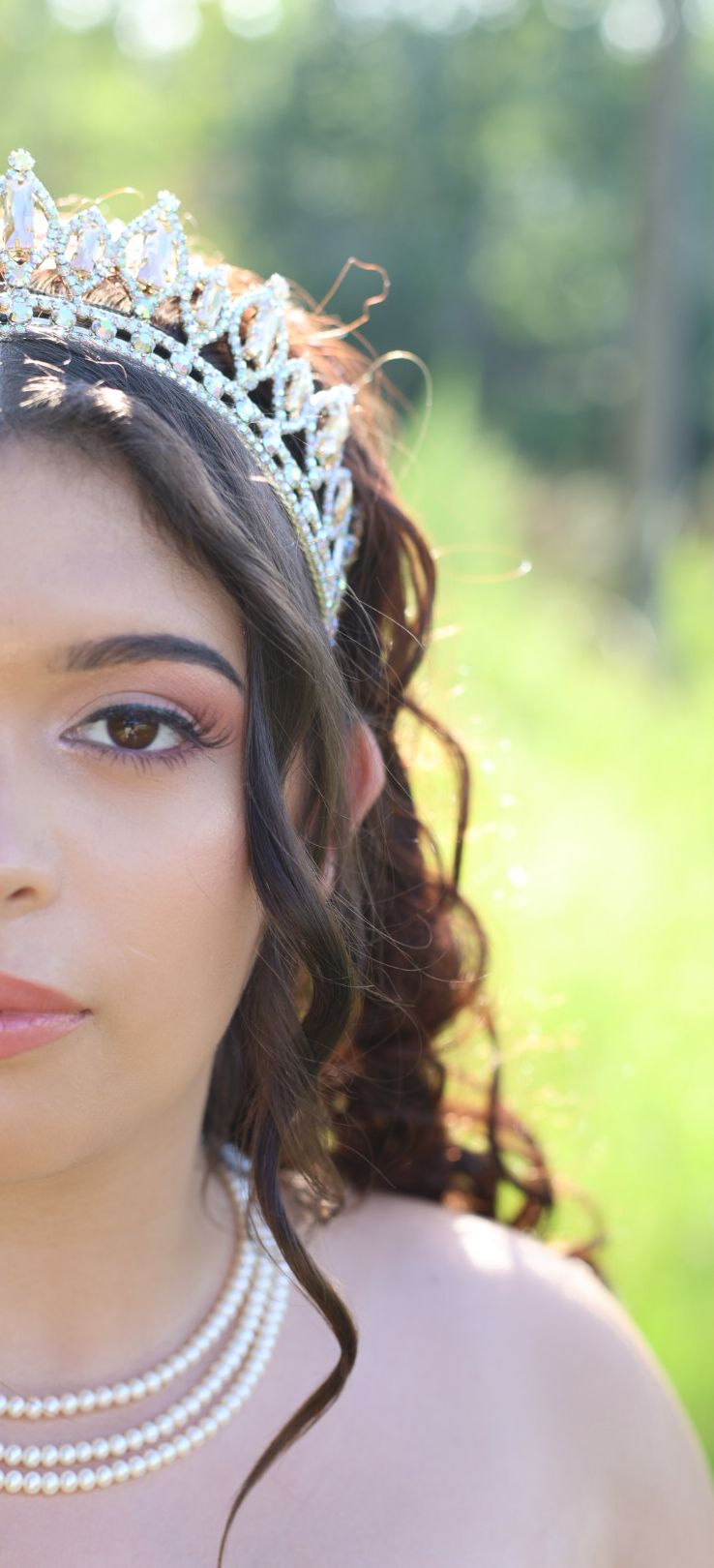 Young woman wearing a sparkling tiara and pearl necklace poses outdoors with soft bokeh green background.
