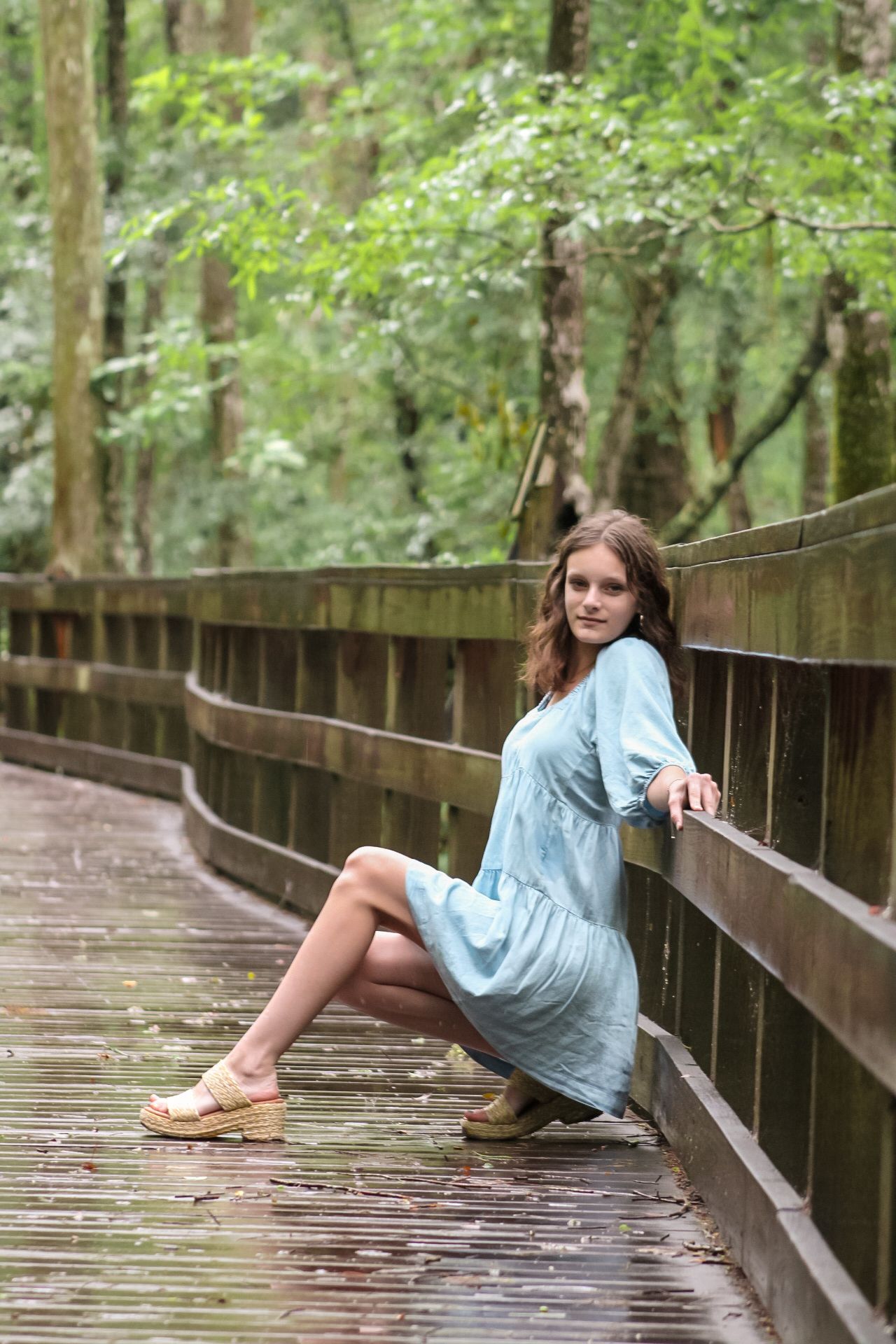 Young woman in light blue dress poses on wooden boardwalk surrounded by lush green forest trees.