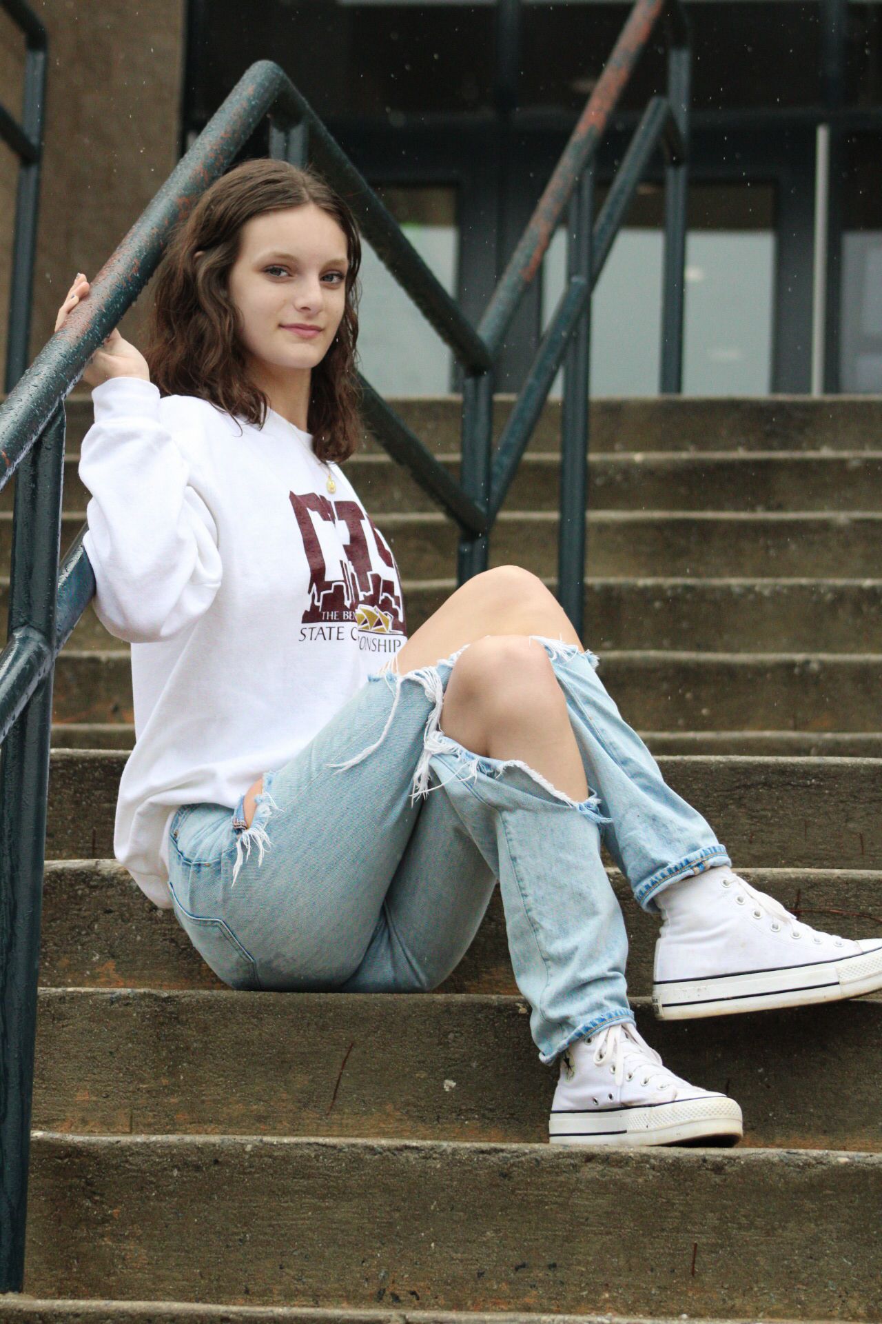 Young woman in white college sweatshirt and ripped jeans sitting on concrete steps wearing white Converse sneakers.