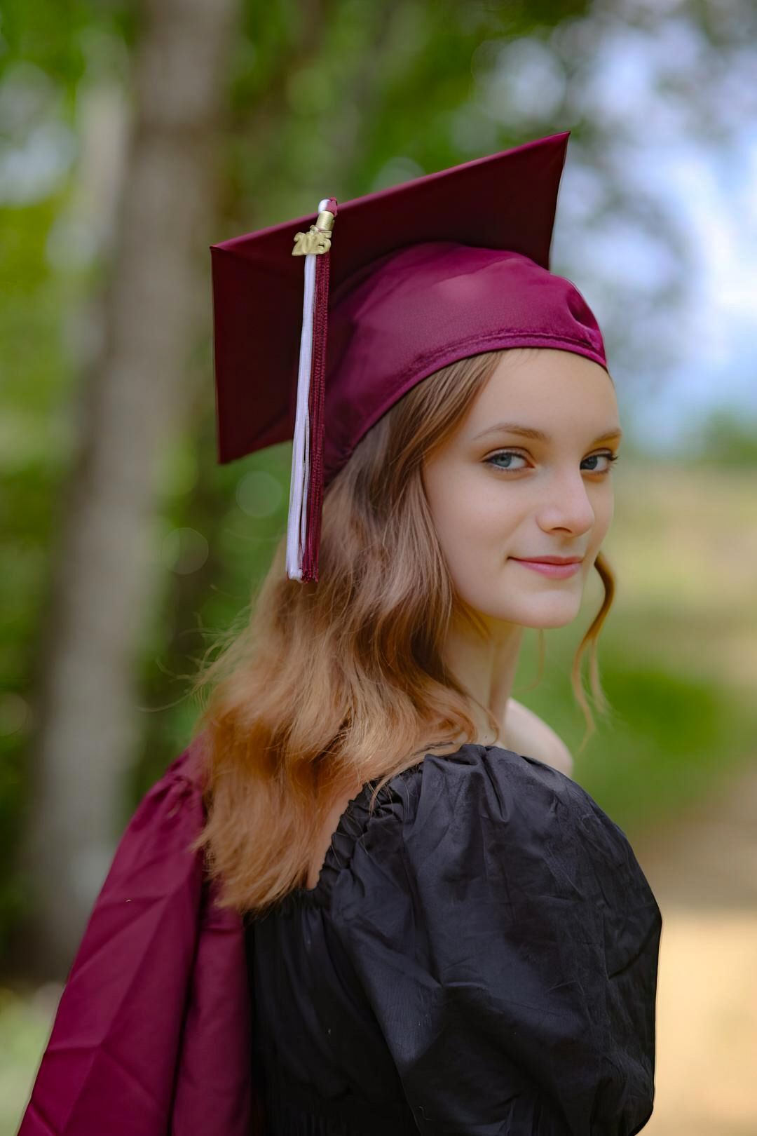 Young woman in maroon graduation cap and gown smiling outdoors among green trees.