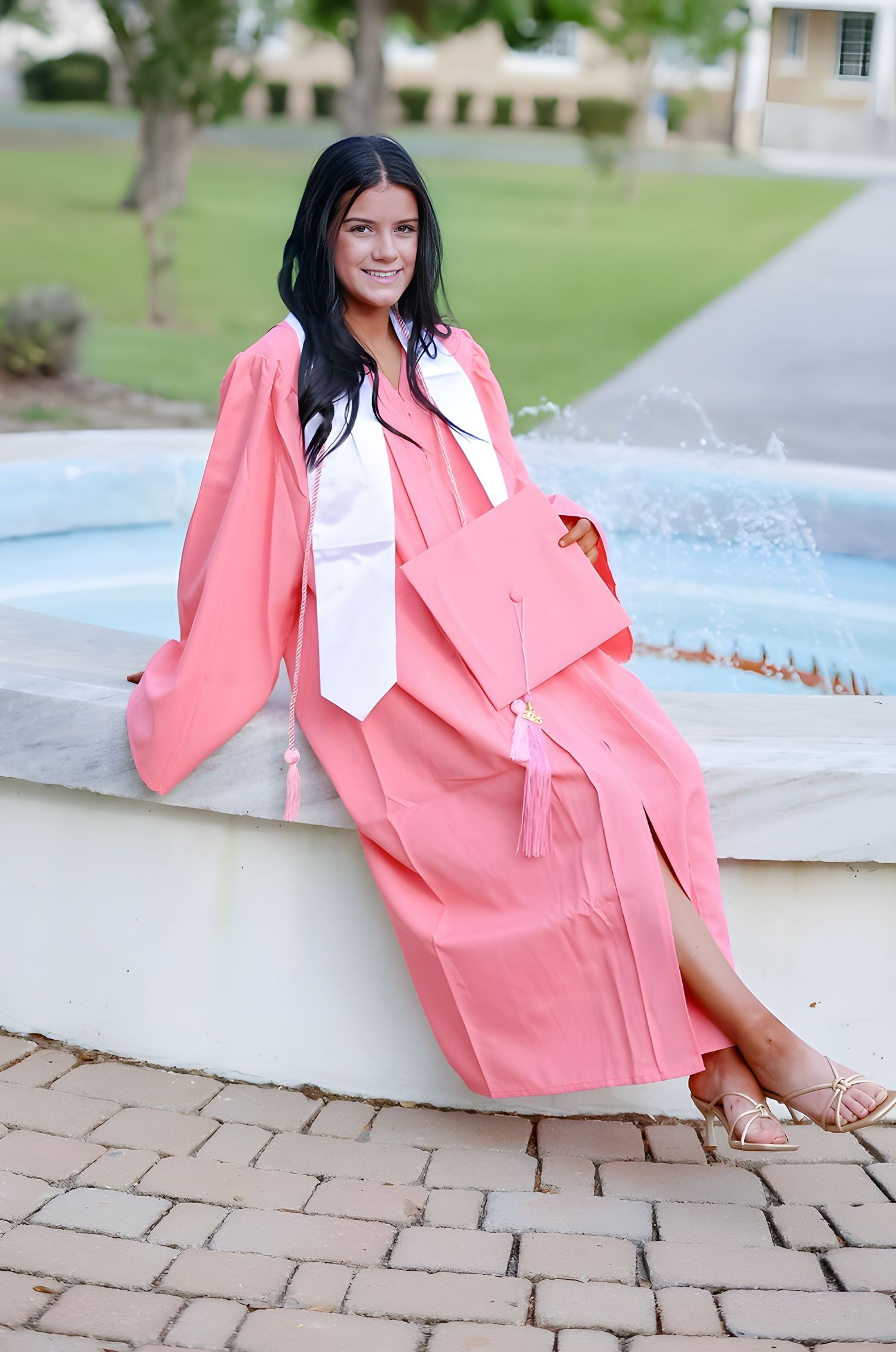 Graduate in coral pink graduation gown and white tie sits gracefully by fountain during outdoor photoshoot.