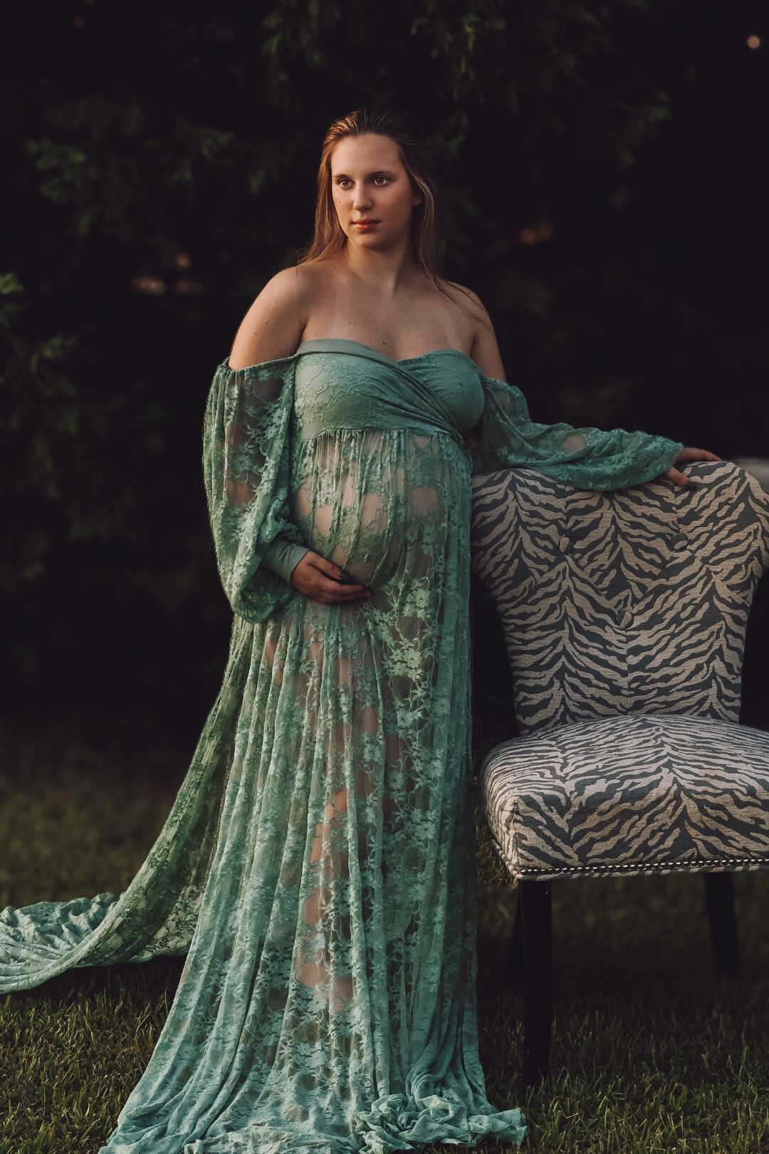 Elegant maternity photo featuring a flowing sage green lace gown and zebra print chair in an outdoor garden setting.