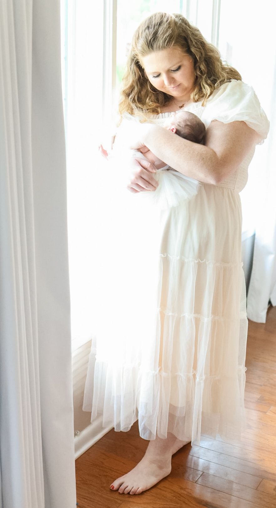 A woman in a flowing white dress stands by a window holding a newborn baby in a tender moment.