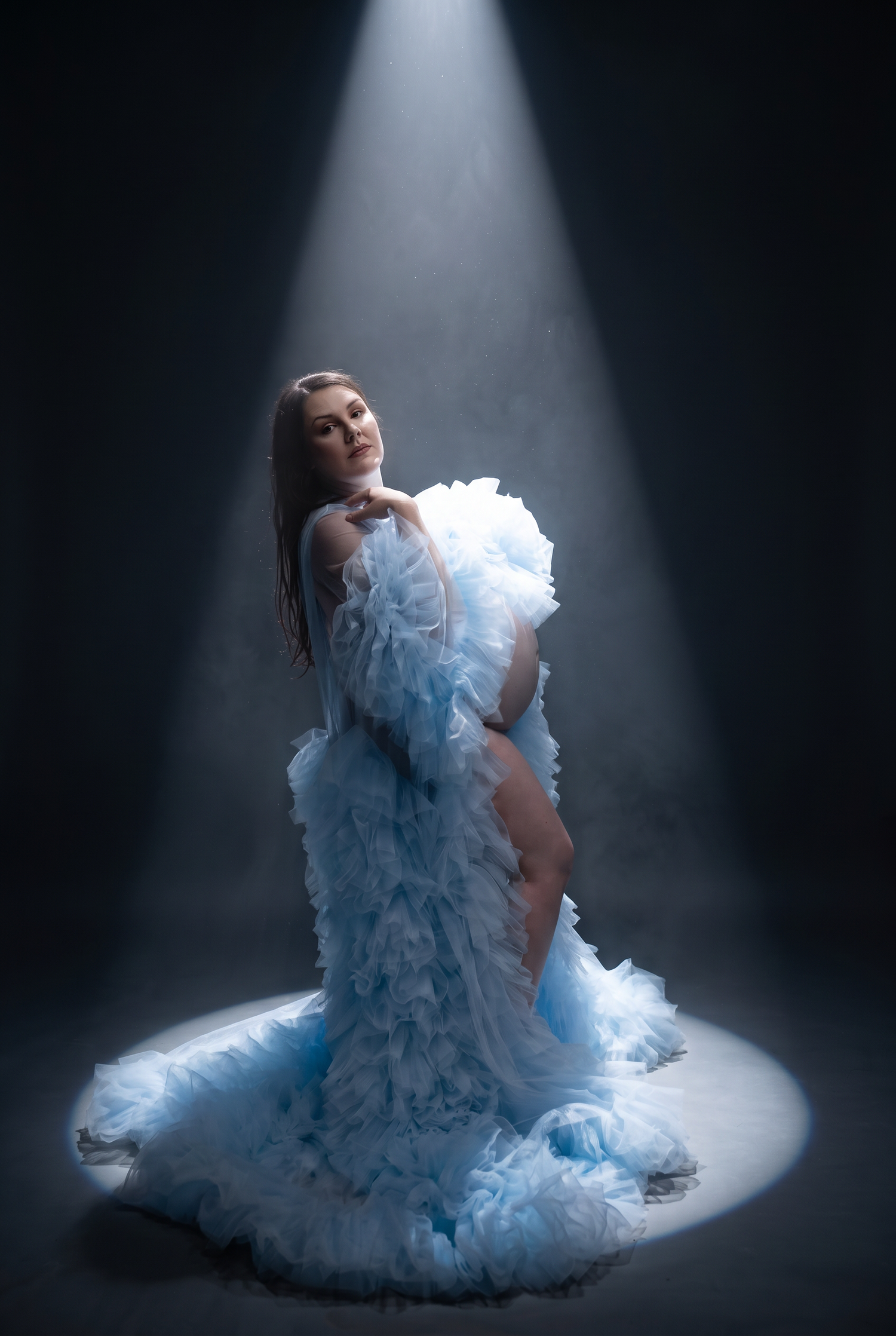 Woman in flowing light blue ruffled gown posed dramatically under a spotlight against a dark smoky background.