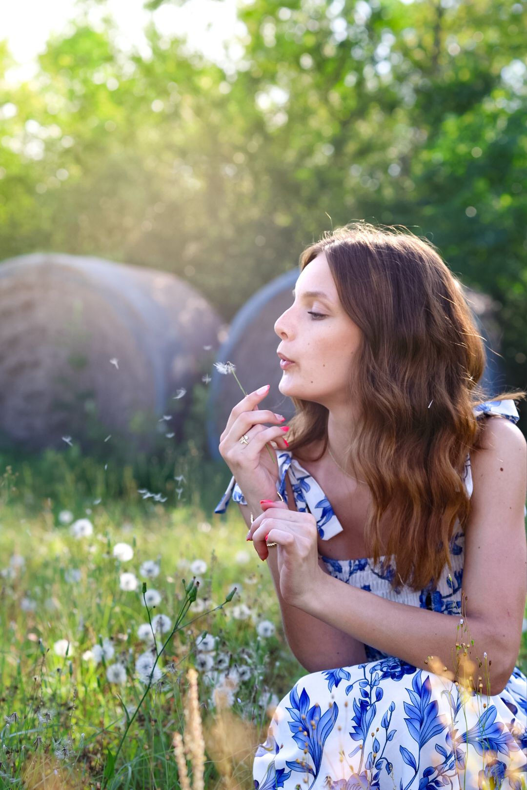 Young woman in blue floral dress blowing dandelion seeds in a sunny garden.
