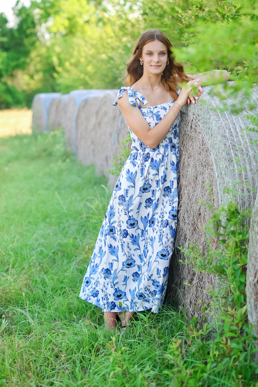 Young woman in blue floral maxi dress leaning against hay bales in a green field.