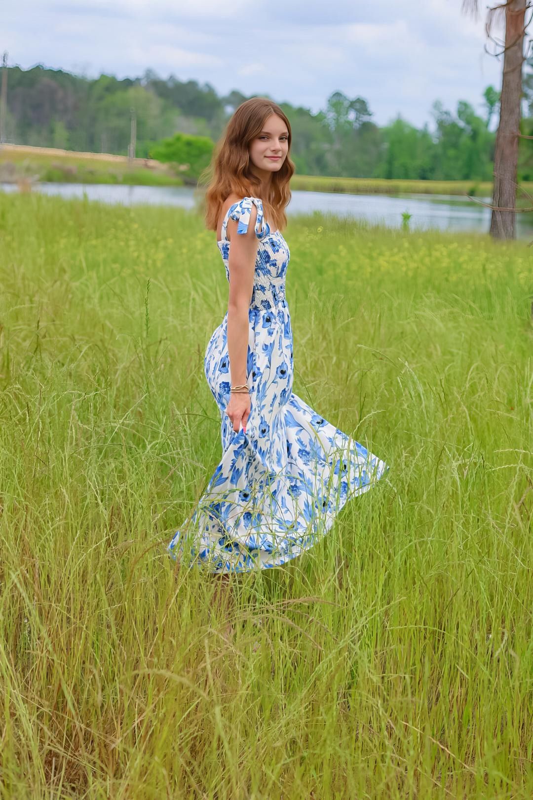 Young woman in blue floral maxi dress standing in tall green grass near a lake on a cloudy day.