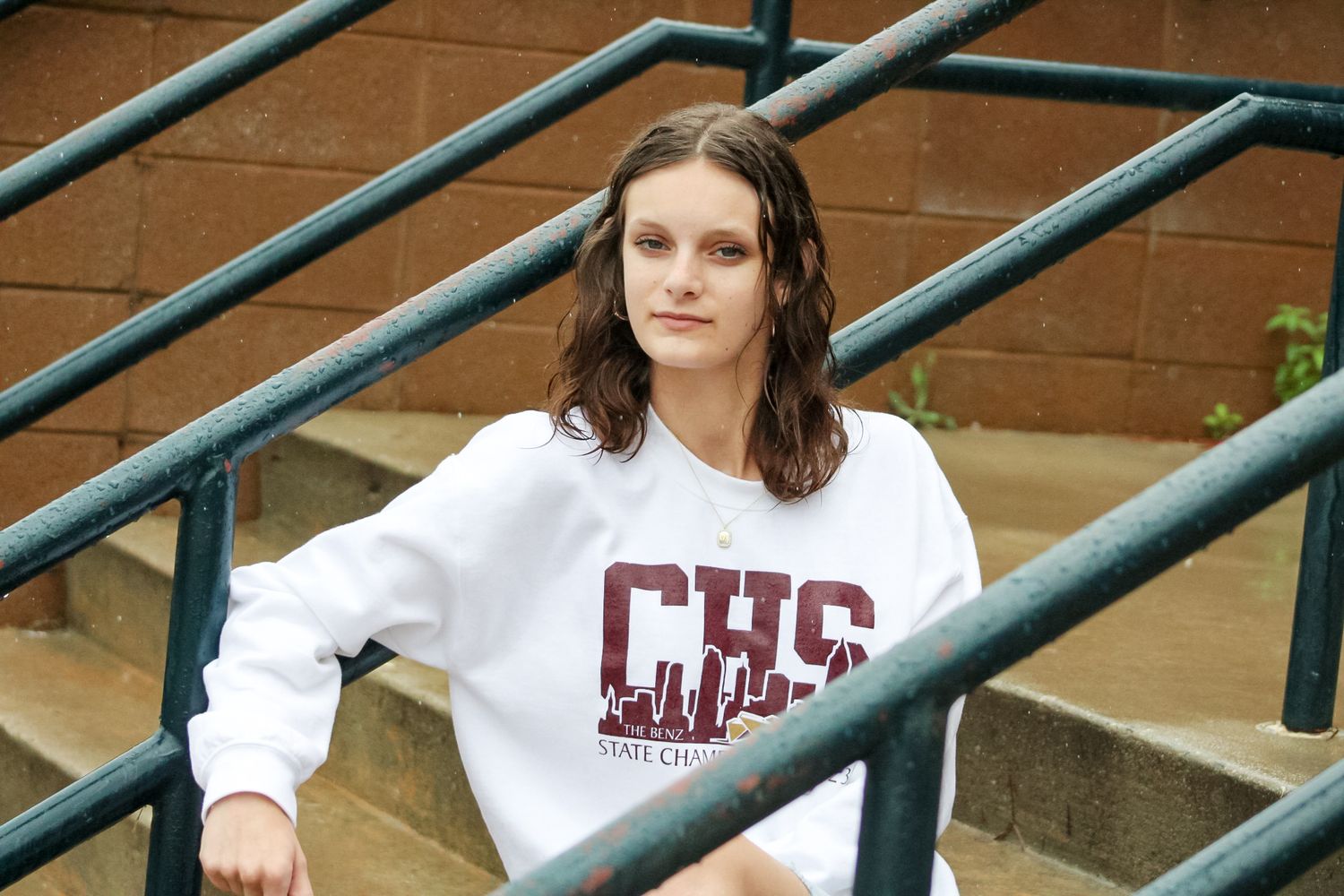 Young woman sitting on bleacher steps wearing a white CHS crewneck sweatshirt outdoors.