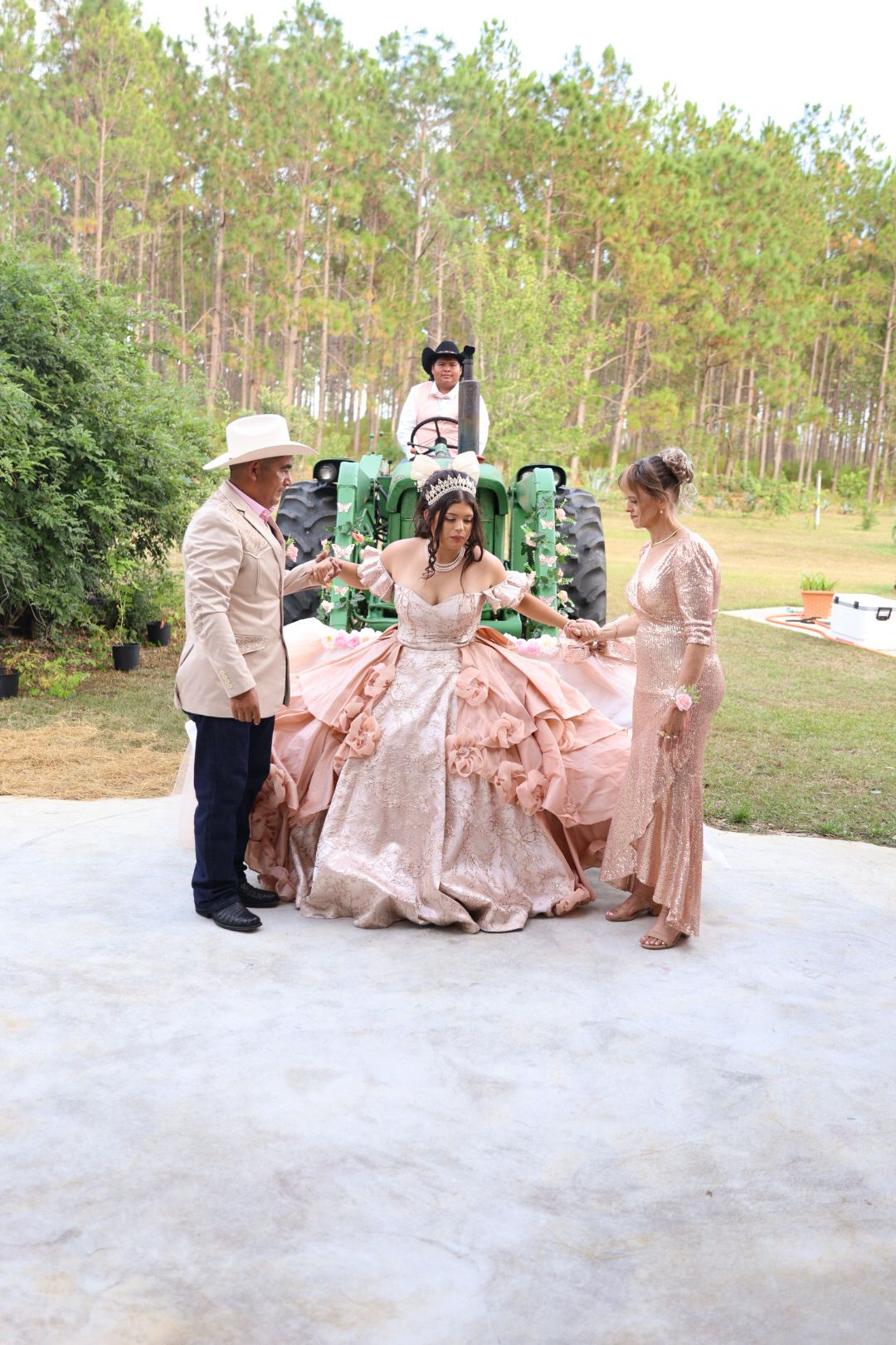 Girl in pink quinceañera gown seated on John Deere tractor surrounded by family in formal attire outdoors.