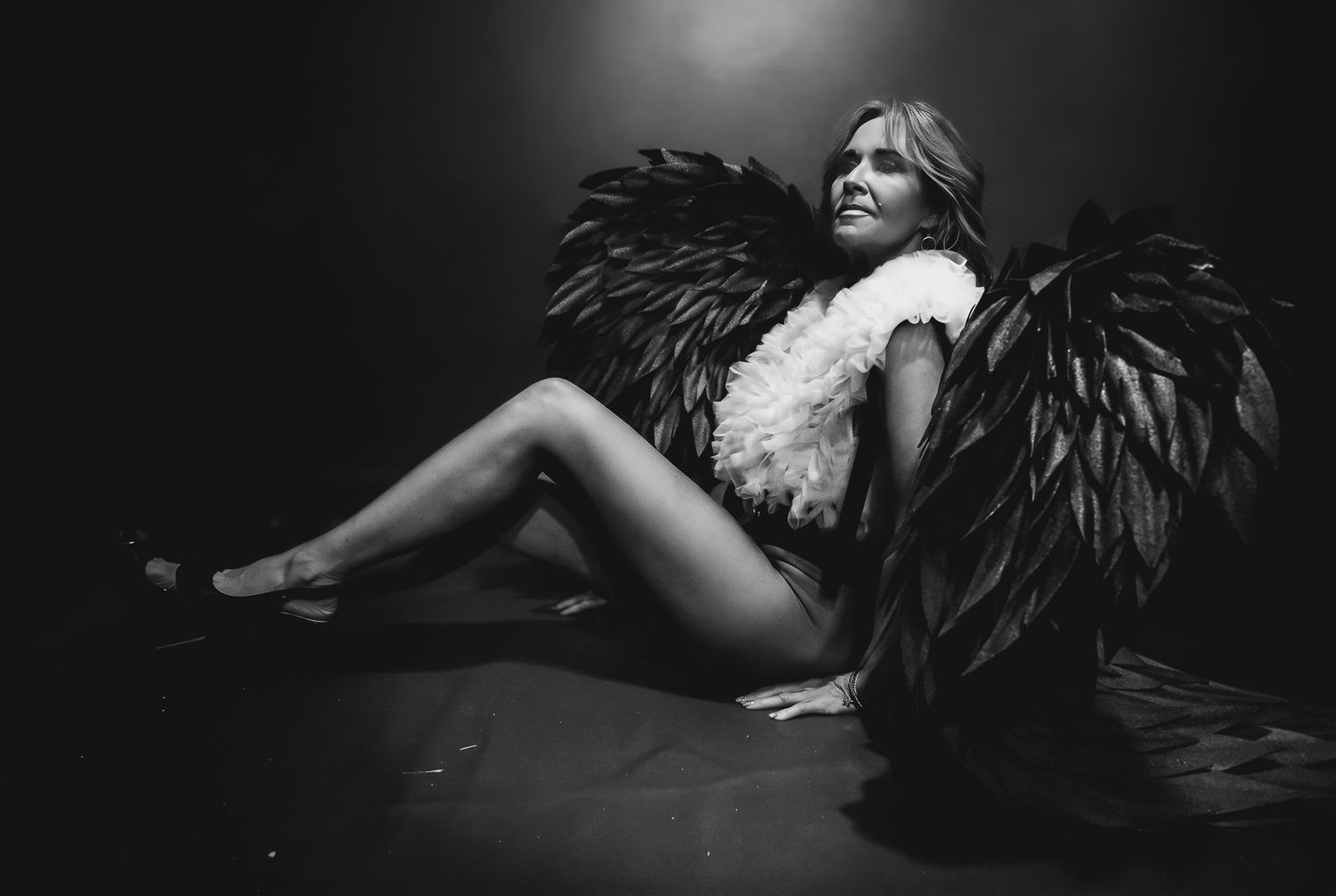 Black and white photo of a woman posing with large dark feathered angel wings in a dramatic studio setting.