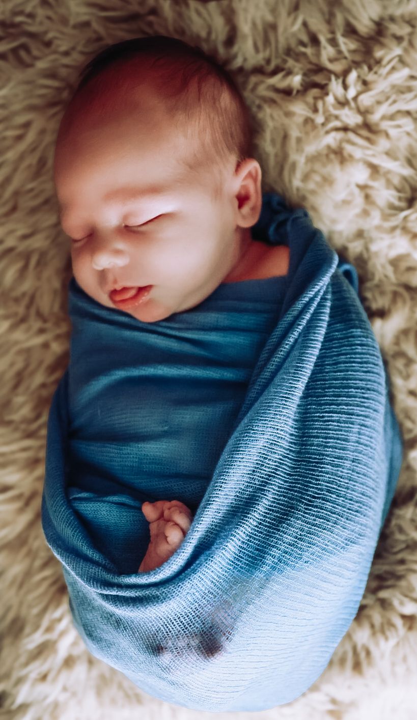 A sleeping newborn baby swaddled in a soft blue gradient knit blanket rests peacefully on fluffy beige fur.