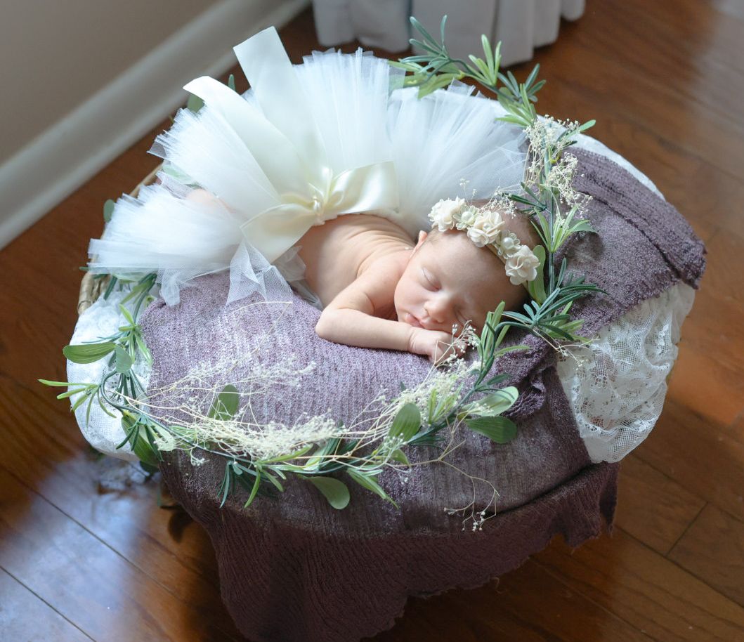 A sleeping newborn wrapped in purple lace fabric with delicate greenery accents rests on a wooden floor.