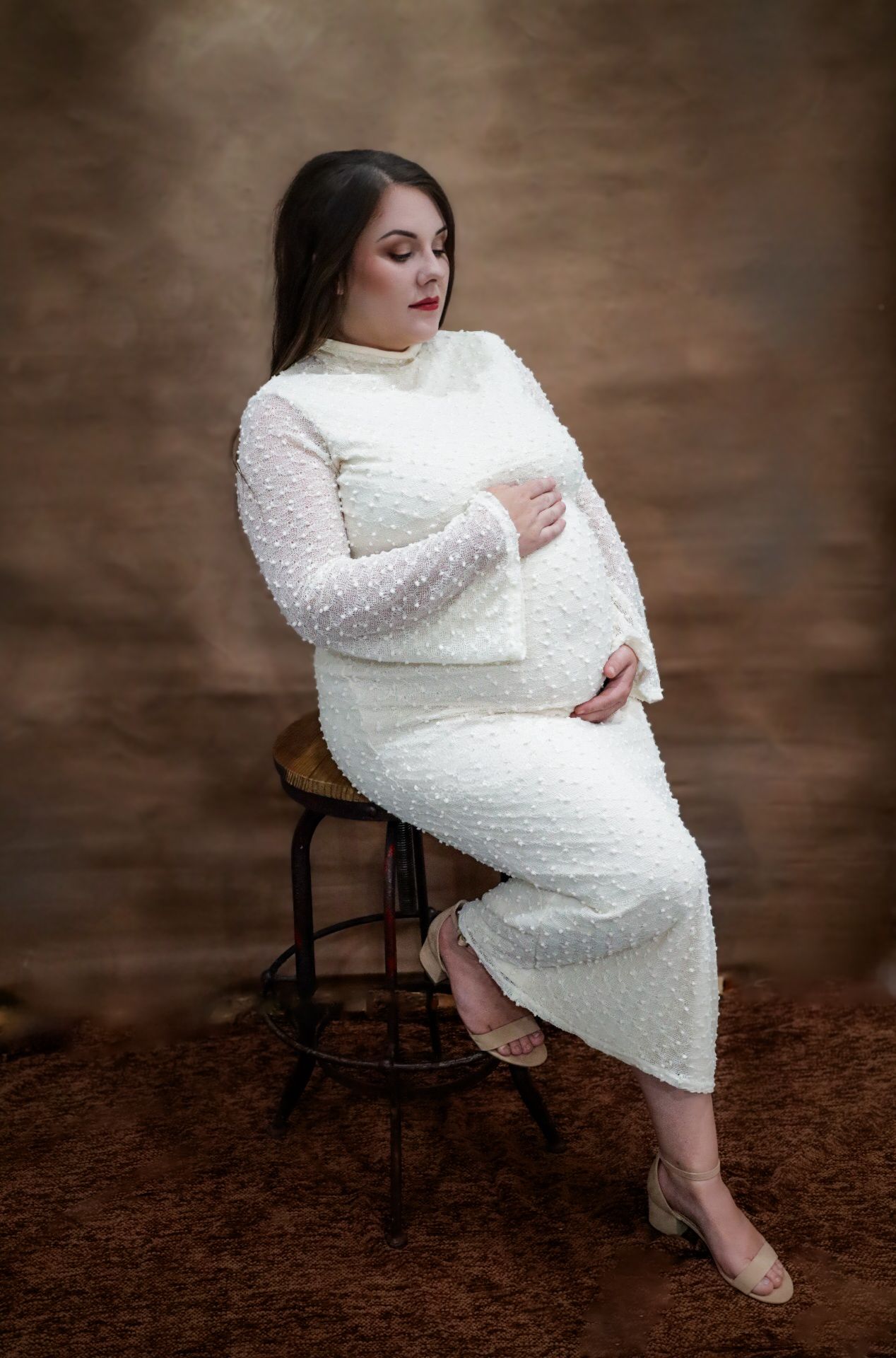 Pregnant woman in white textured long-sleeve gown sitting on stool, cradling baby bump in professional studio portrait.