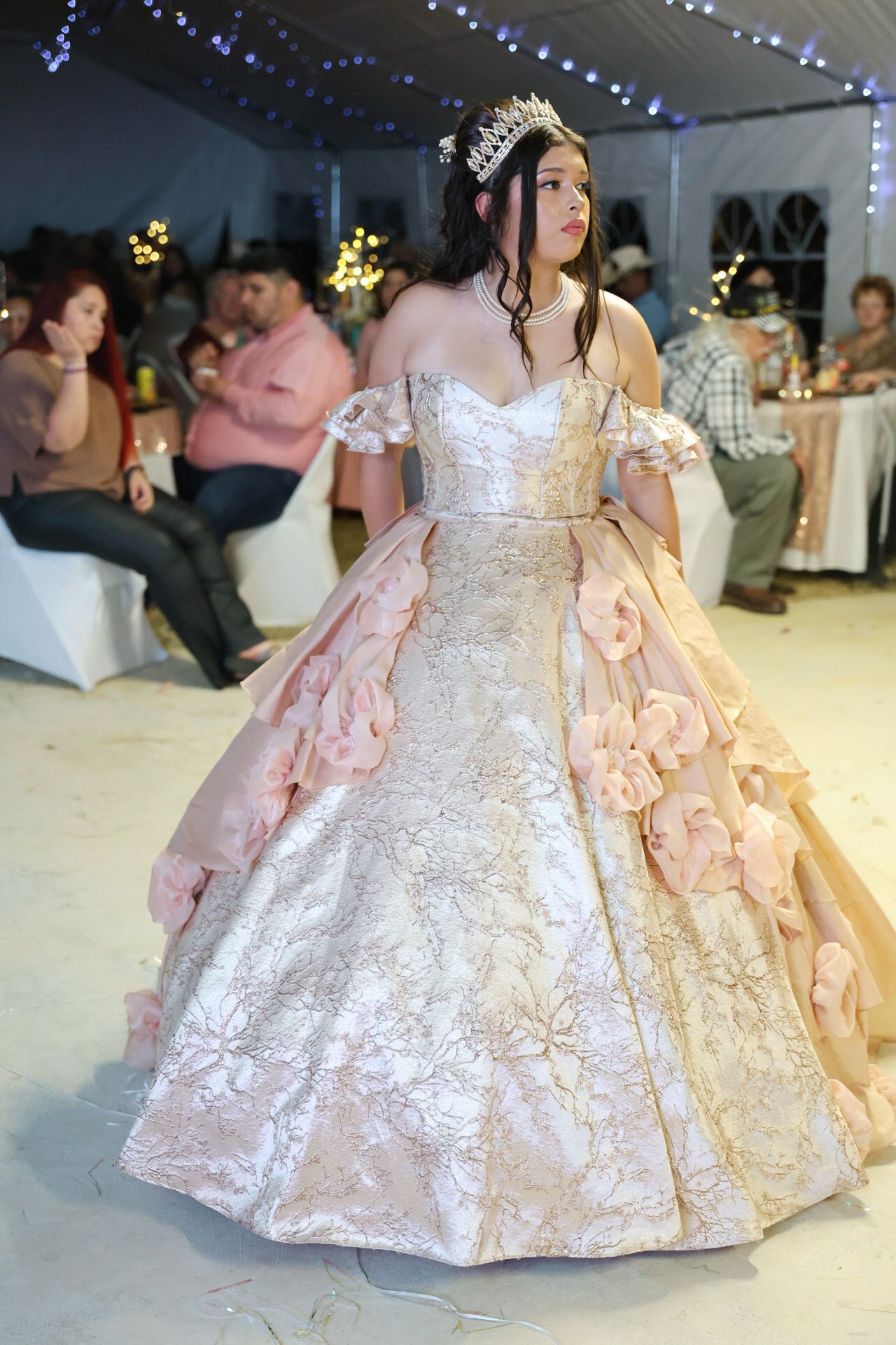 Young woman in elegant pink ball gown with floral details and tiara at a quinceañera celebration.