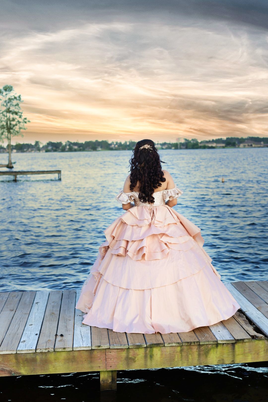 Young woman in pink ball gown stands on wooden dock overlooking a serene lake at sunset.