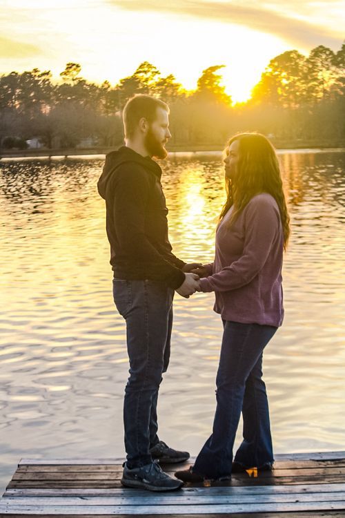 Couple holding hands on a wooden dock at golden sunset with a reflective lake and trees in the background.