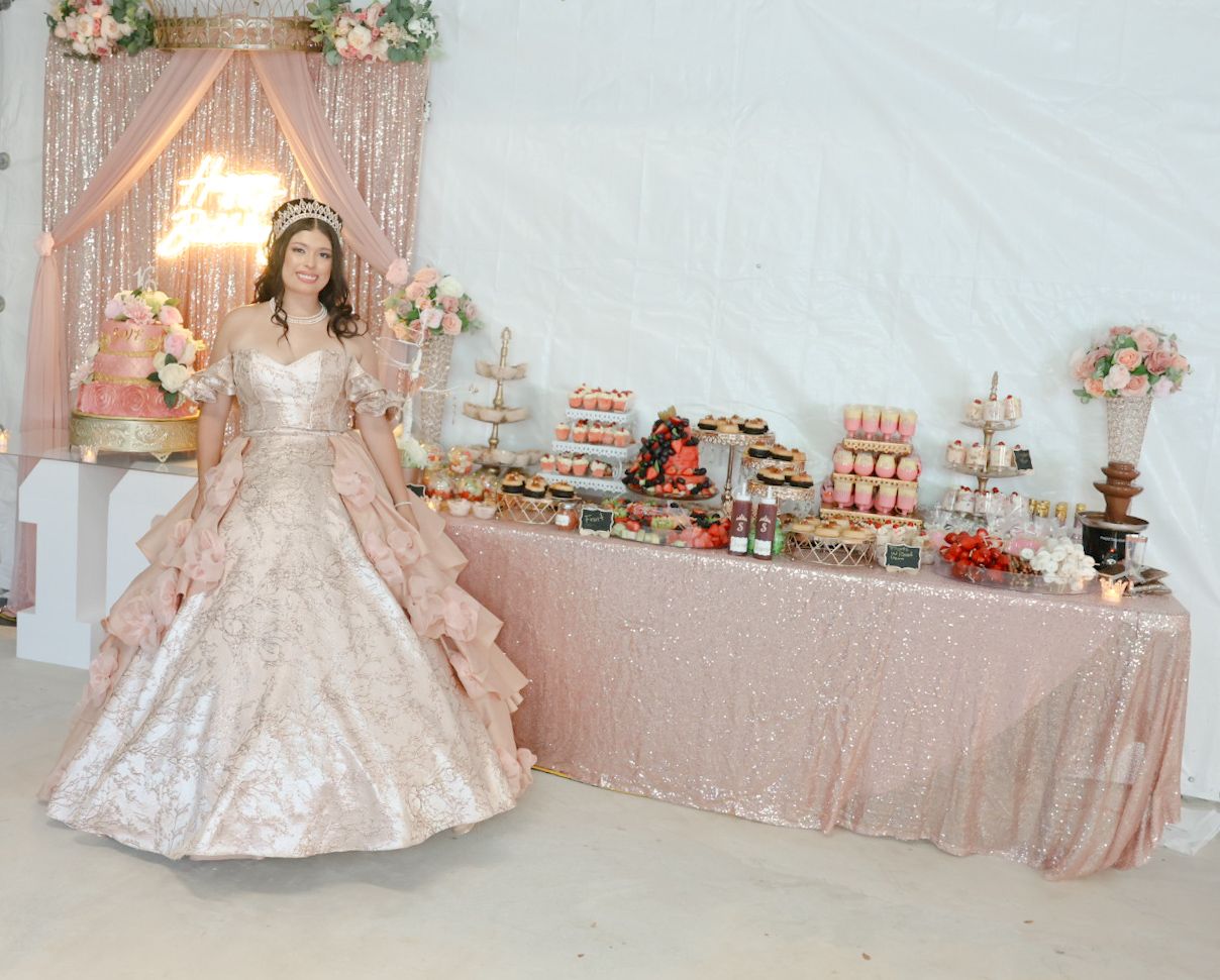 Young woman in elegant rose gold quinceañera gown stands beside a decorated dessert table at a celebration.
