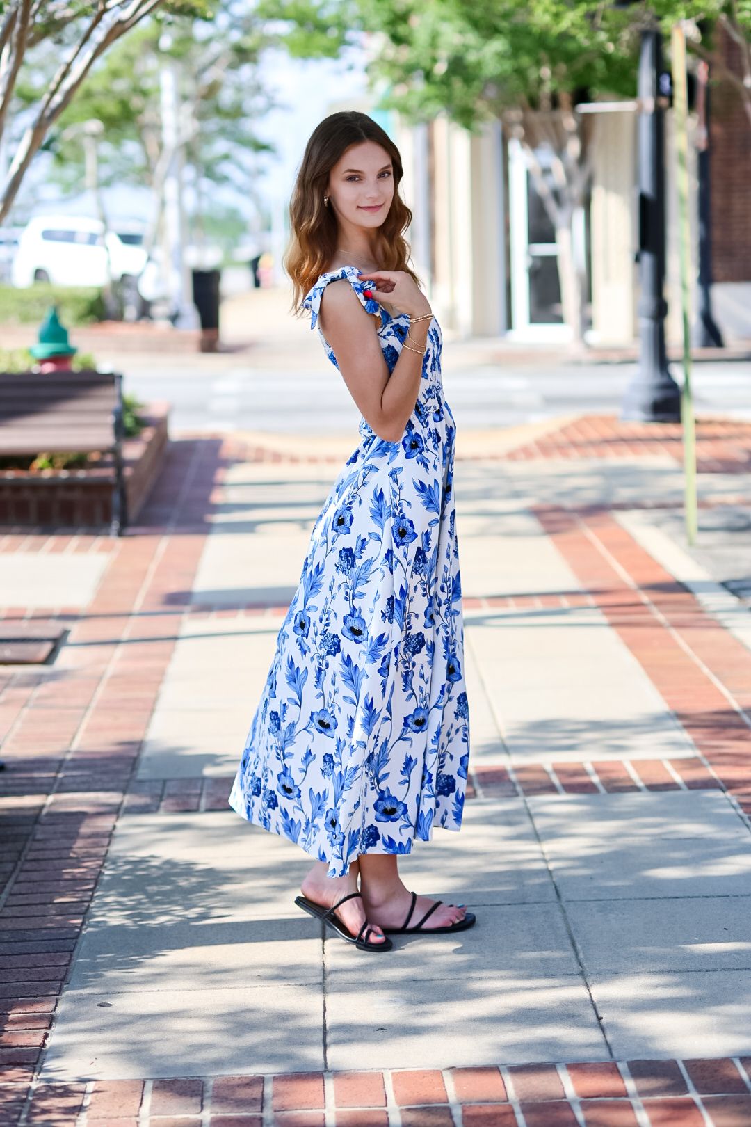 Young woman in blue floral maxi dress glancing back on brick downtown sidewalk.