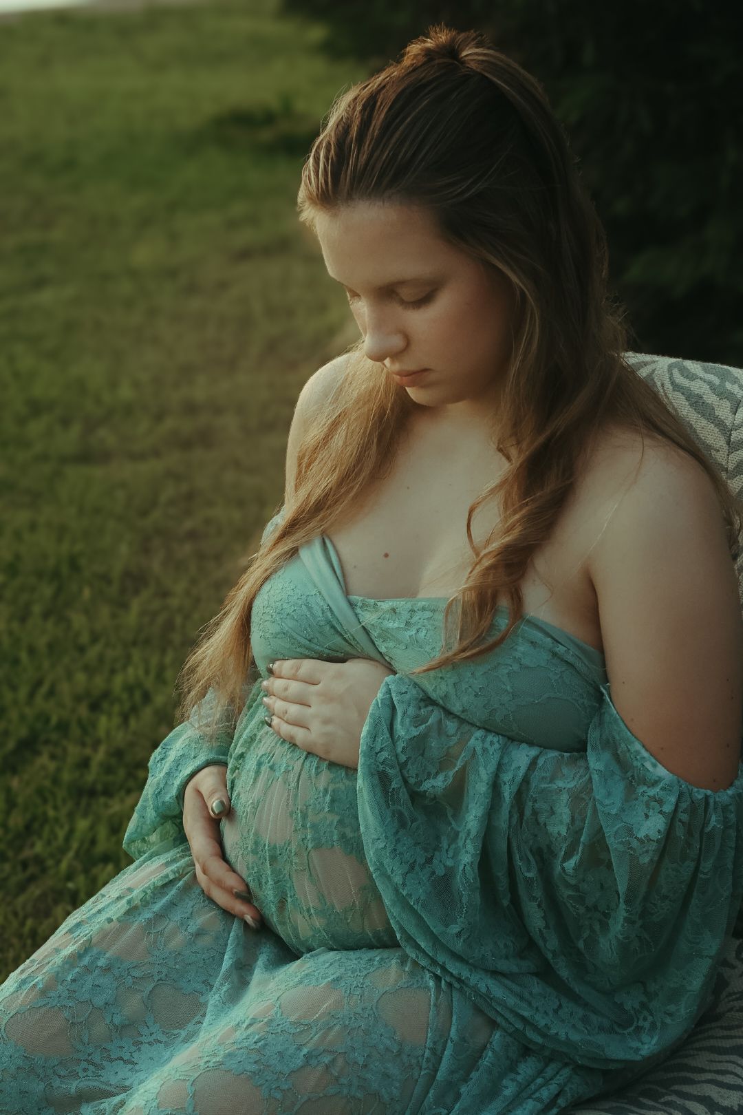 A woman in a turquoise lace maternity dress poses thoughtfully in a grassy outdoor setting during golden hour.