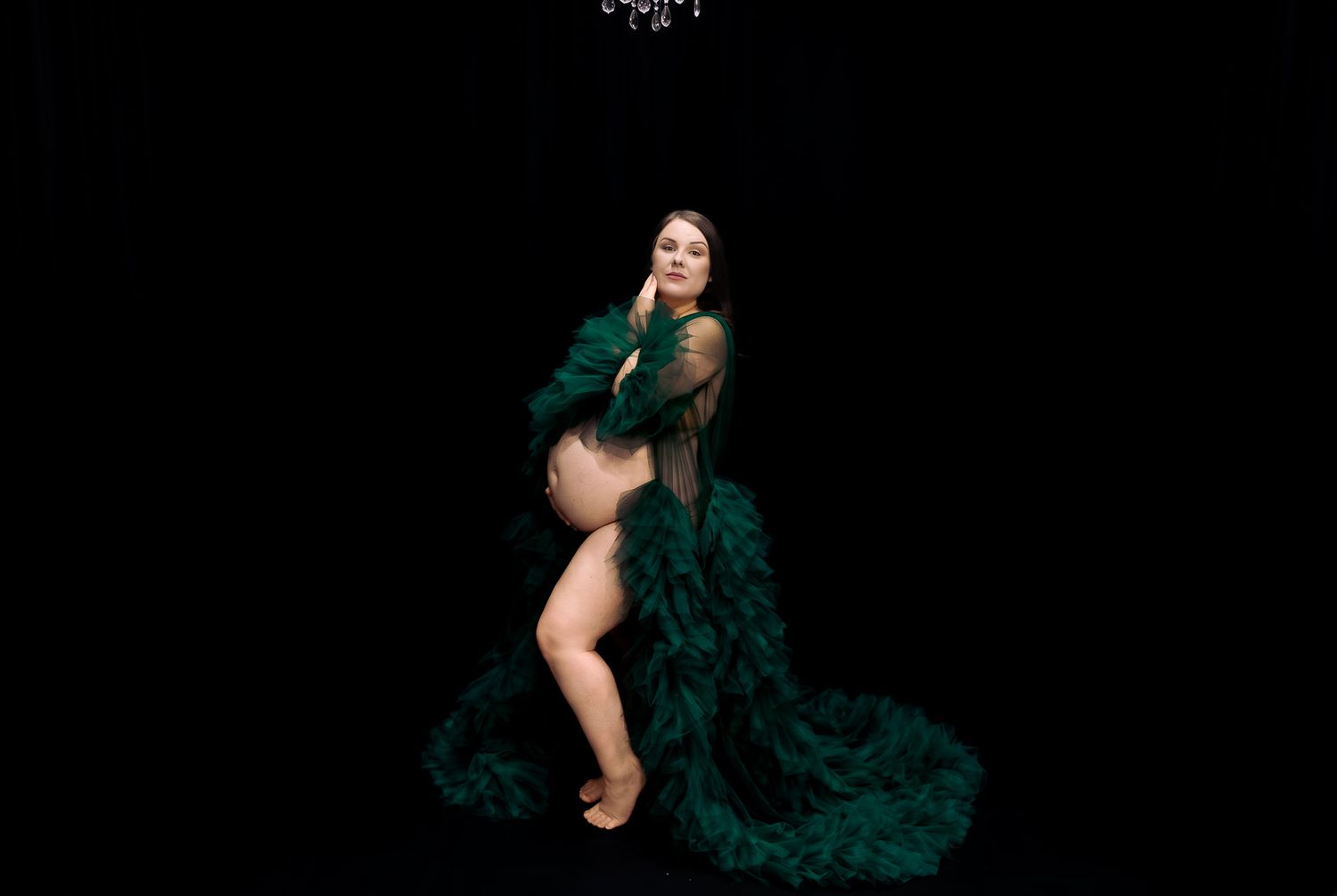 Pregnant woman posing in elegant dark green feathered robe gown against black background in maternity photo shoot.