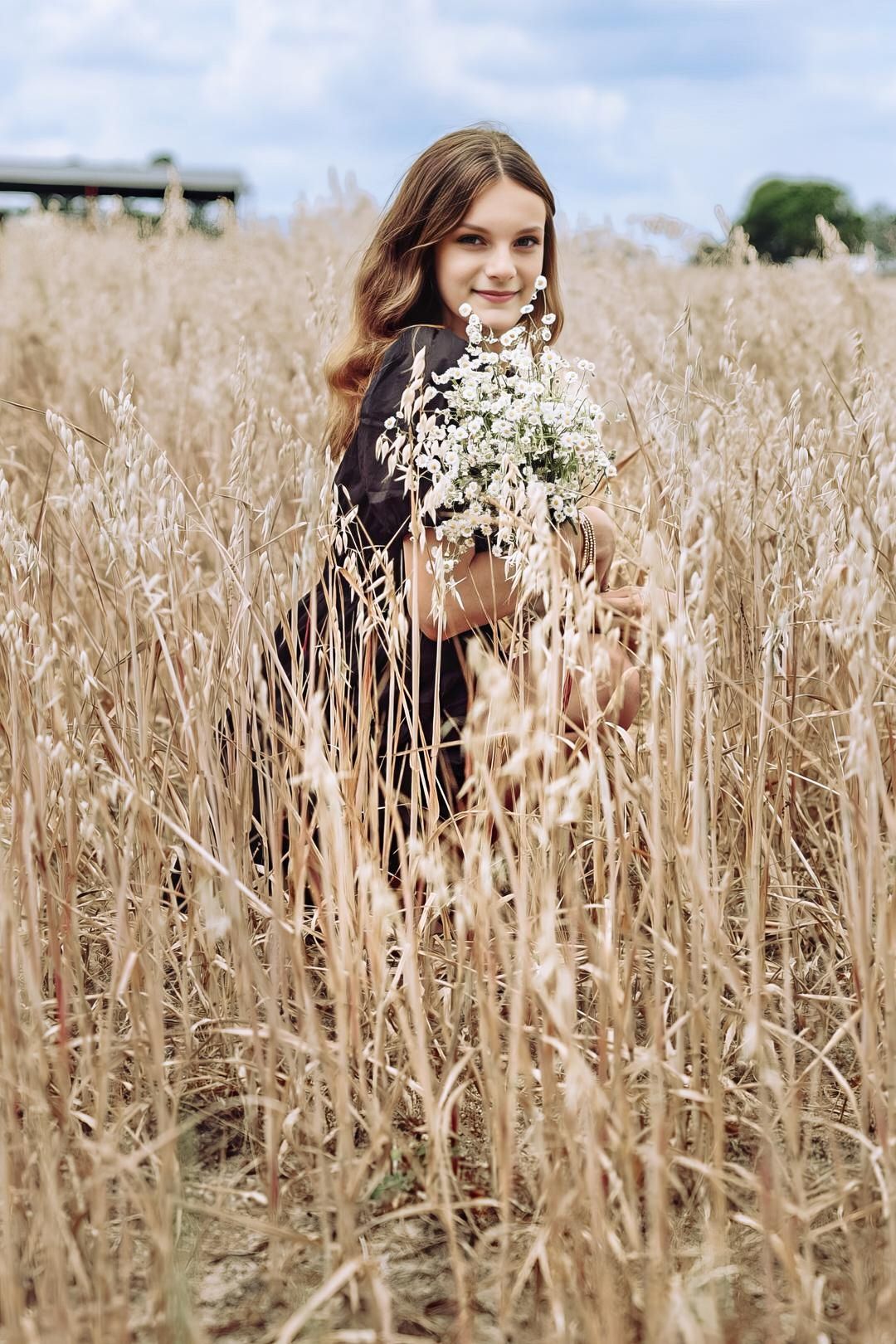 Young woman in dark dress holding wildflowers while standing in golden wheat field.