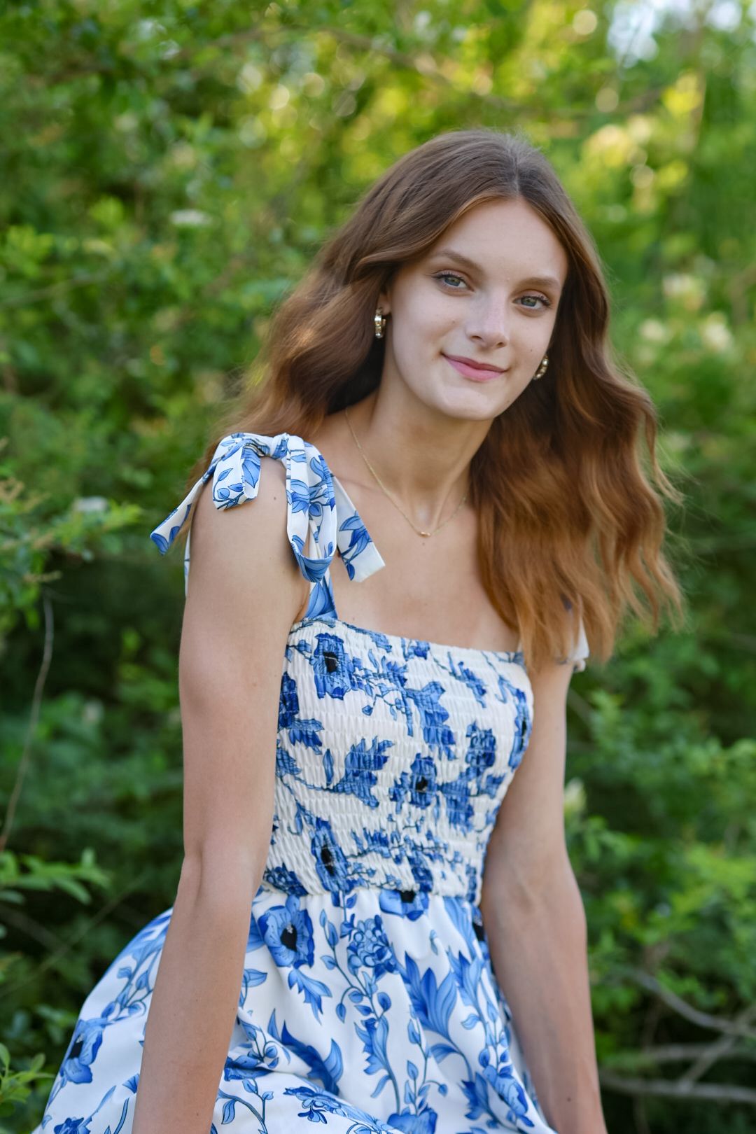 Young woman in blue floral sundress smiling outdoors surrounded by green foliage.