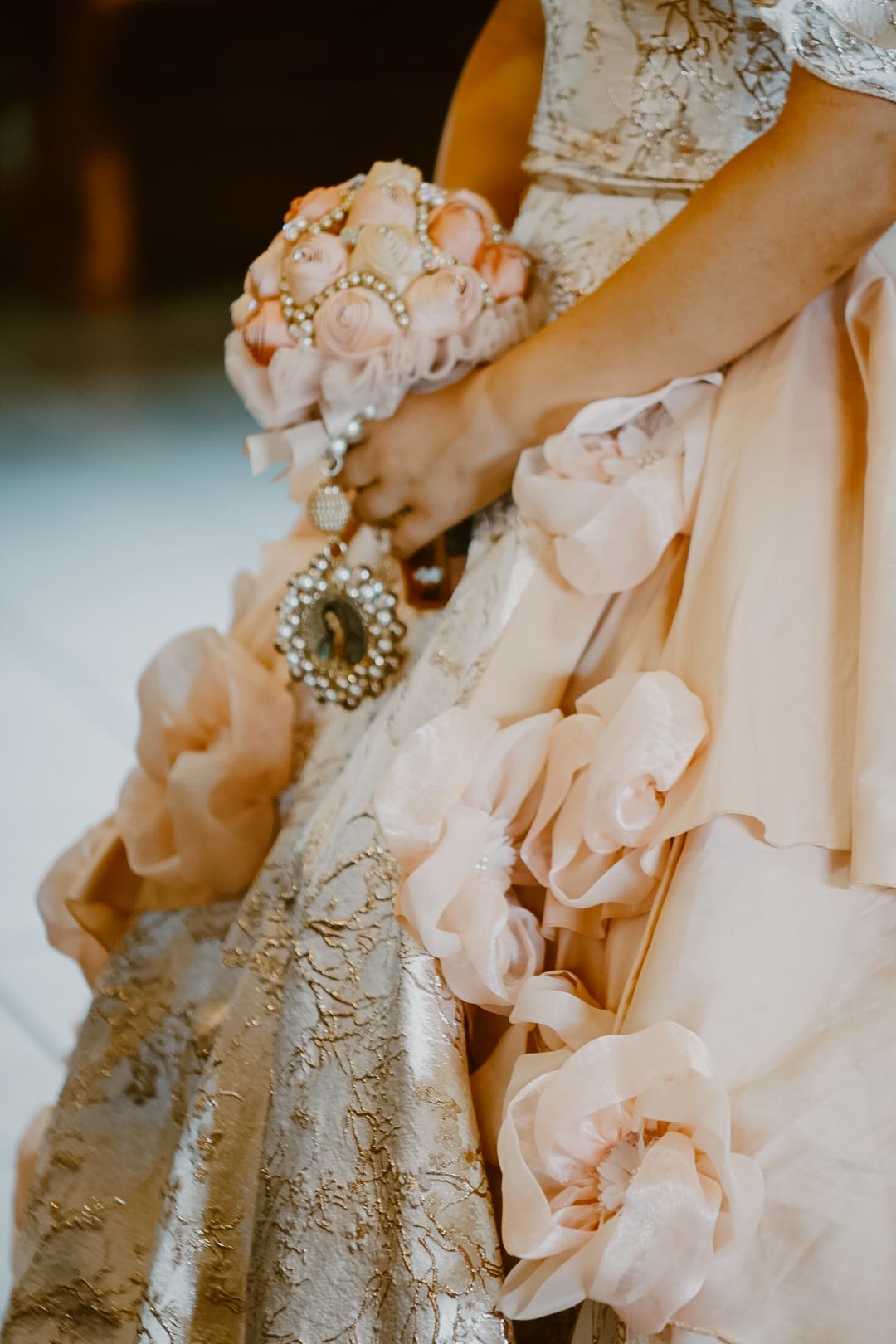 Bride holding a pearl bouquet in a blush pink ruffled wedding gown with gold floral embroidery details.