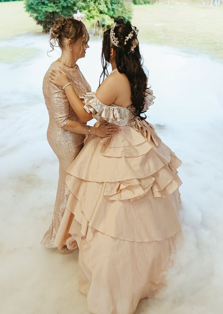 Two women in elegant blush gowns share a tender dance moment surrounded by dreamy fog at a formal event.