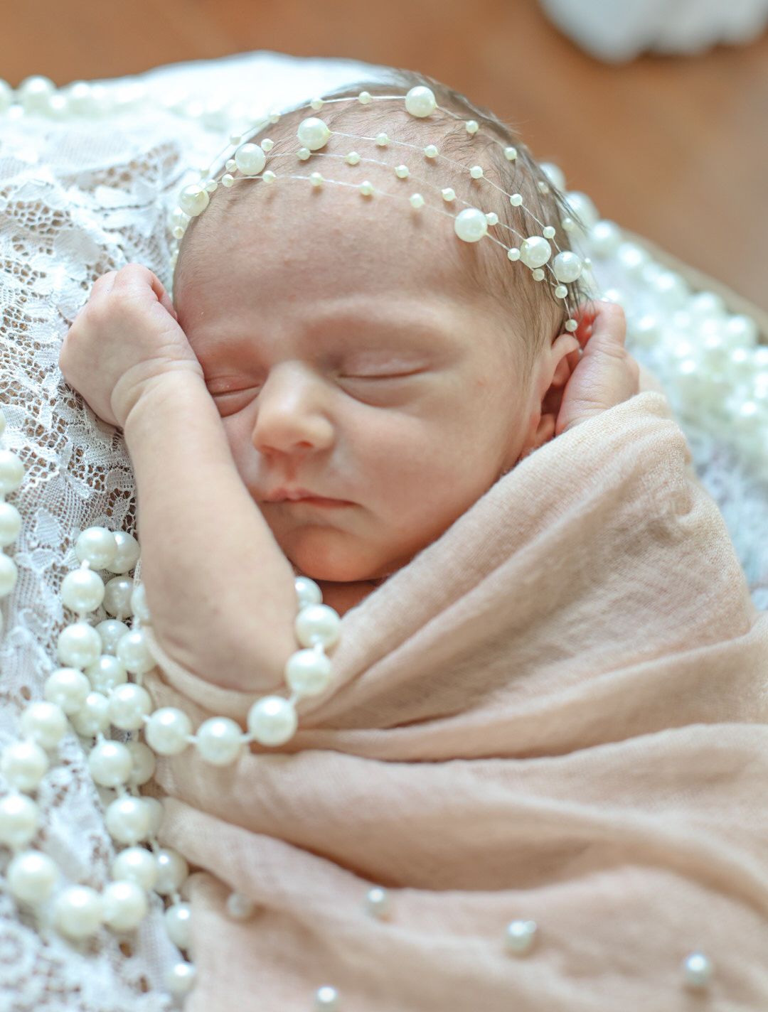 A sleeping newborn baby wrapped in soft pink fabric wearing a delicate pearl headband.