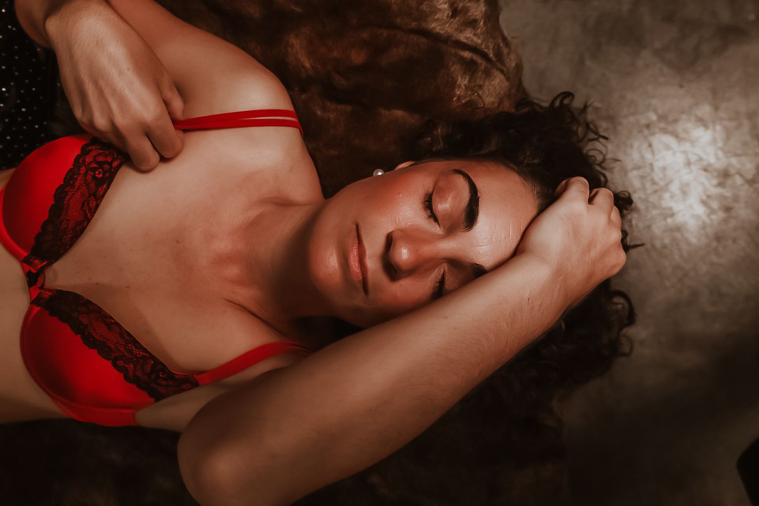 A woman in red lingerie lies on the floor with eyes closed, her curly hair spread around her in a moody, artistic boudoir photo.