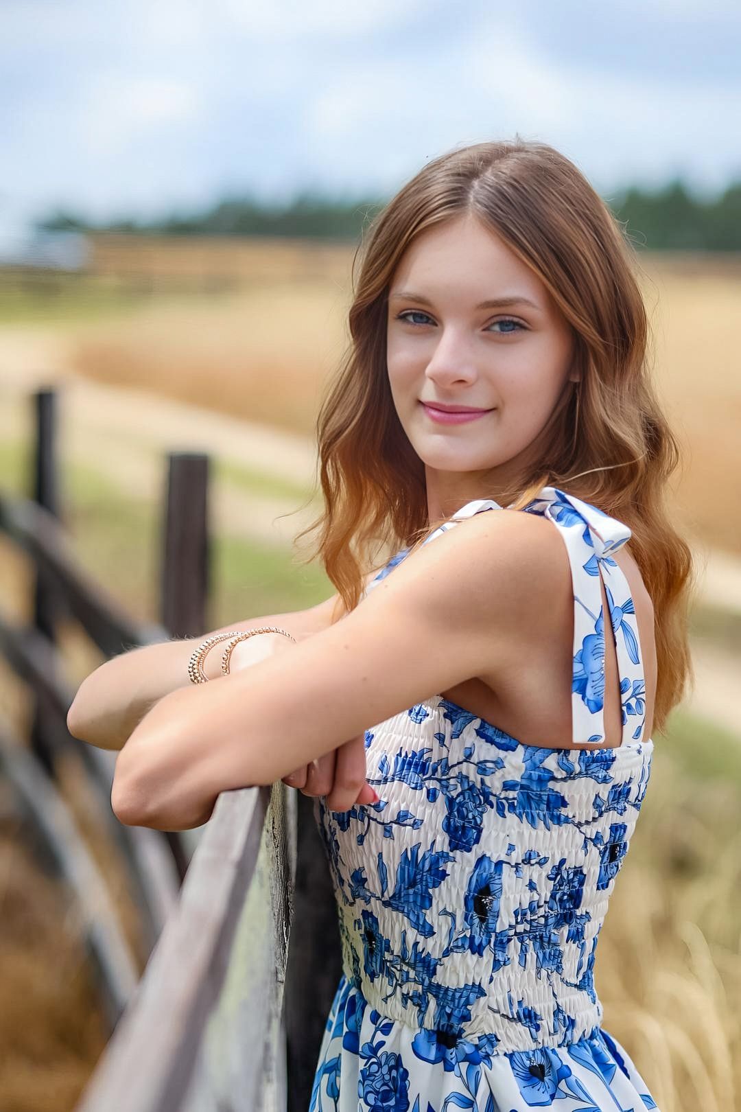 A person in a blue and white floral sundress leans against a wooden fence in a sunny rural field.