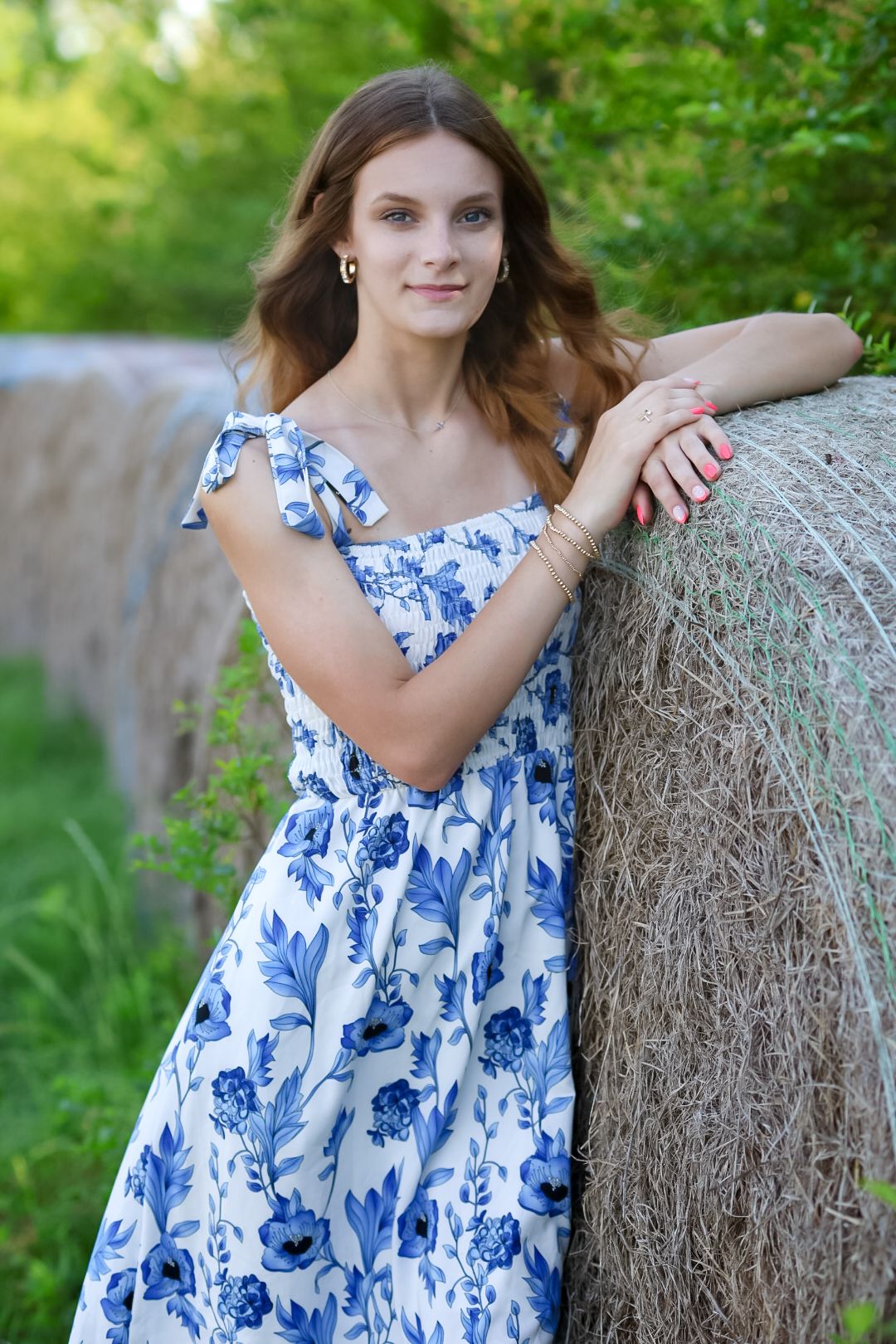 Young woman in blue floral dress leaning on hay bale outdoors.