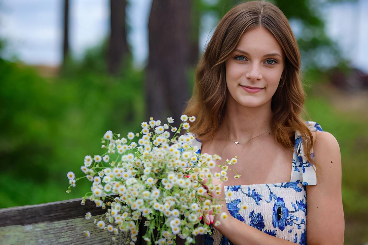 Young woman in blue floral dress holding white wildflowers bouquet, smiling outdoors in a garden setting.