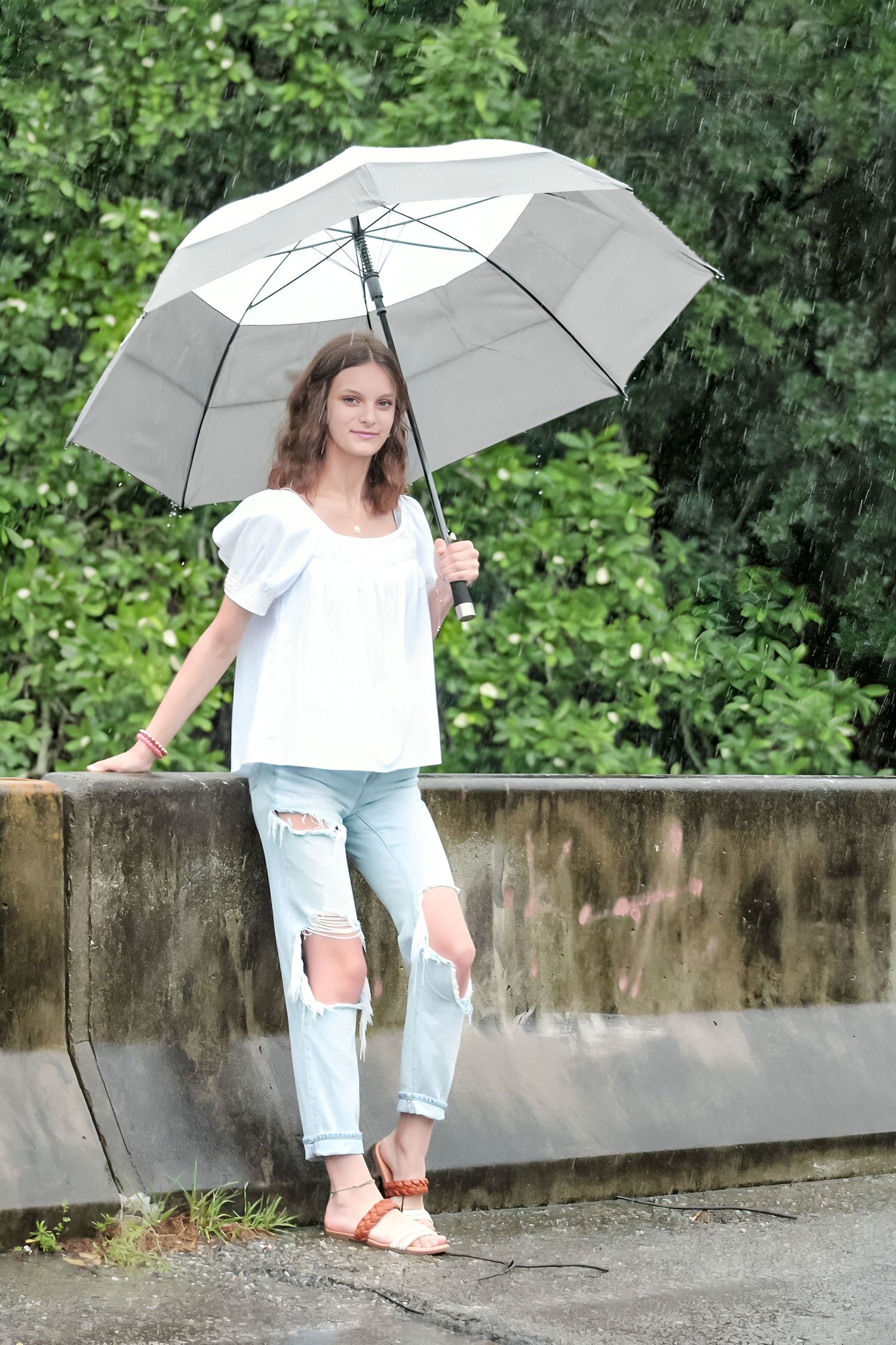 Young woman in white top and ripped jeans holding a gray umbrella in the rain outdoors.