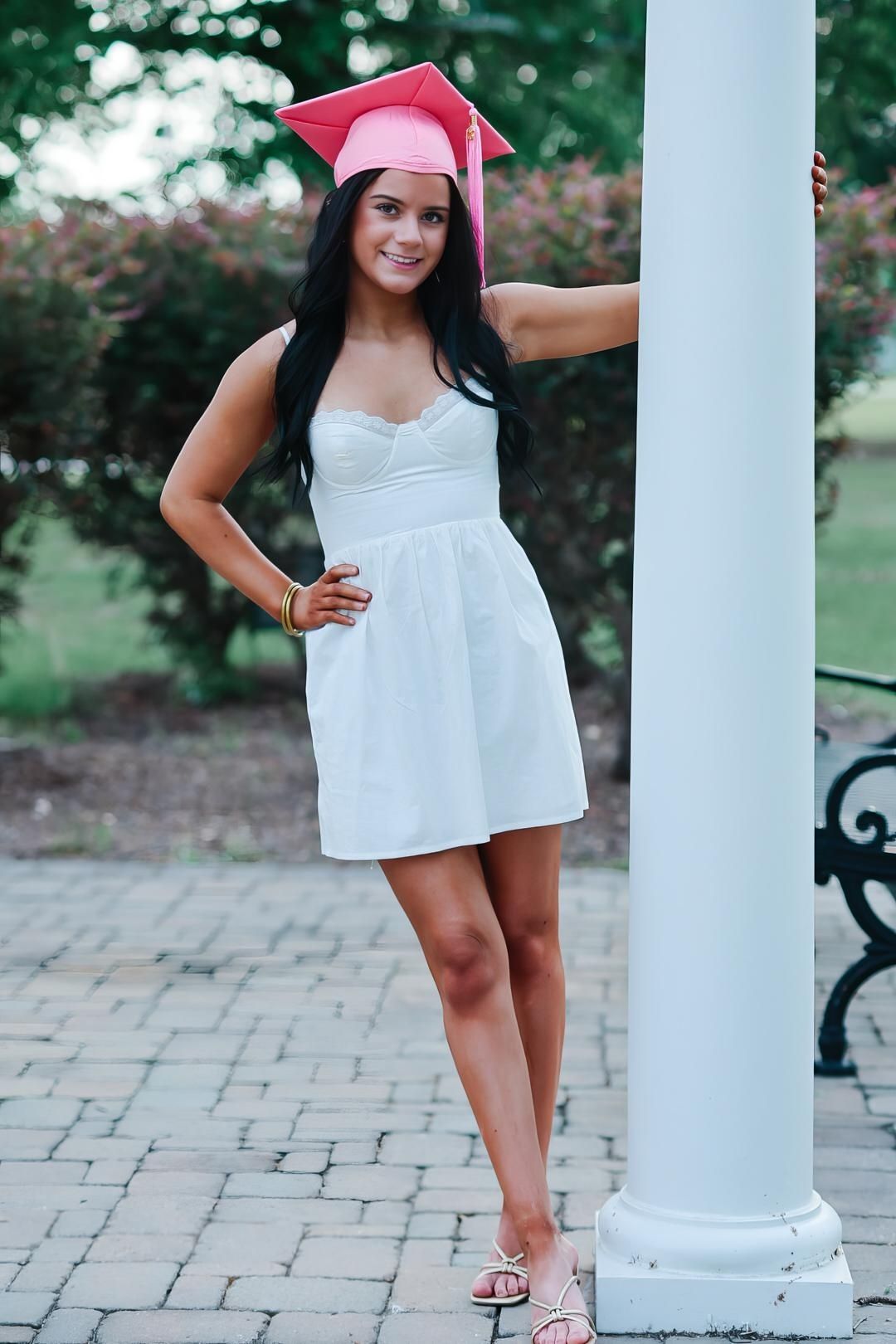 Graduate in white summer dress and pink cap poses by decorative column on brick patio during outdoor photo session.
