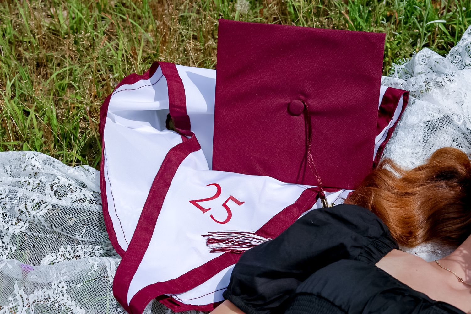 Person lying on grass with maroon graduation cap and white stole numbered 25 for a graduation photo.