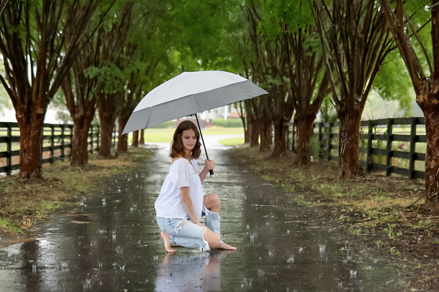Woman kneeling on wet road holding silver umbrella in rain, surrounded by tree-lined path with wooden fence.