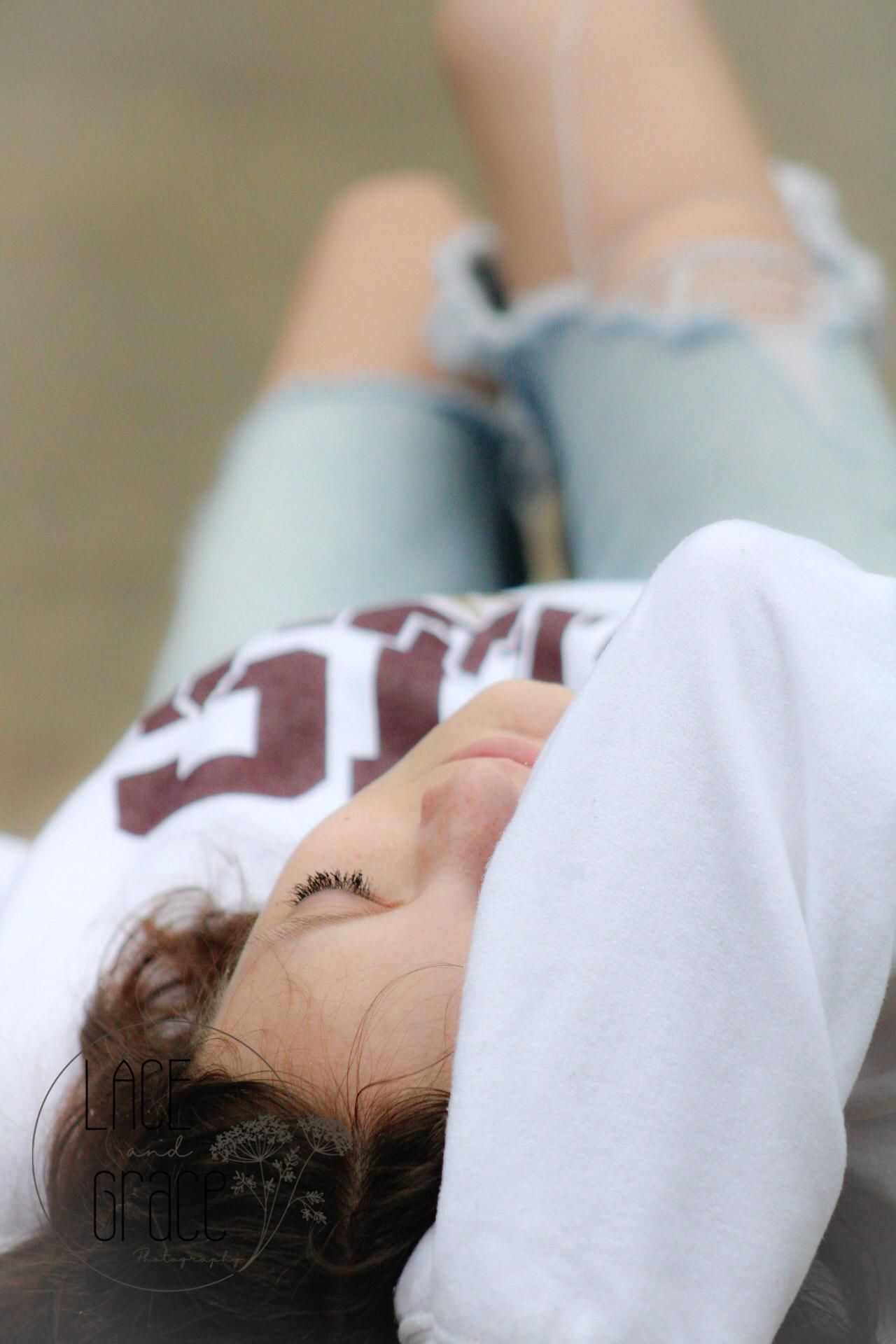 Young woman lying down outdoors, eyes closed, wearing white CHS sweatshirt and ripped jeans.