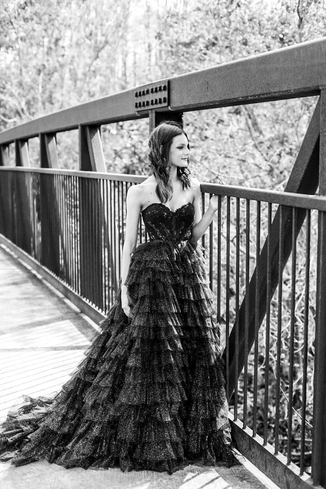 Black and white photo of a woman in a strapless ruffled ball gown standing on a wooden bridge.