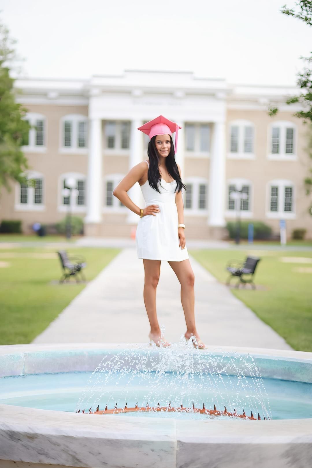 A graduate in a white dress and pink cap stands on a fountain ledge at a college campus with a classic building behind.