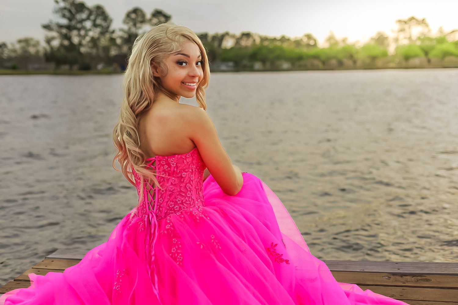 A person in a vibrant hot pink ball gown poses on a wooden dock at sunset near the water.